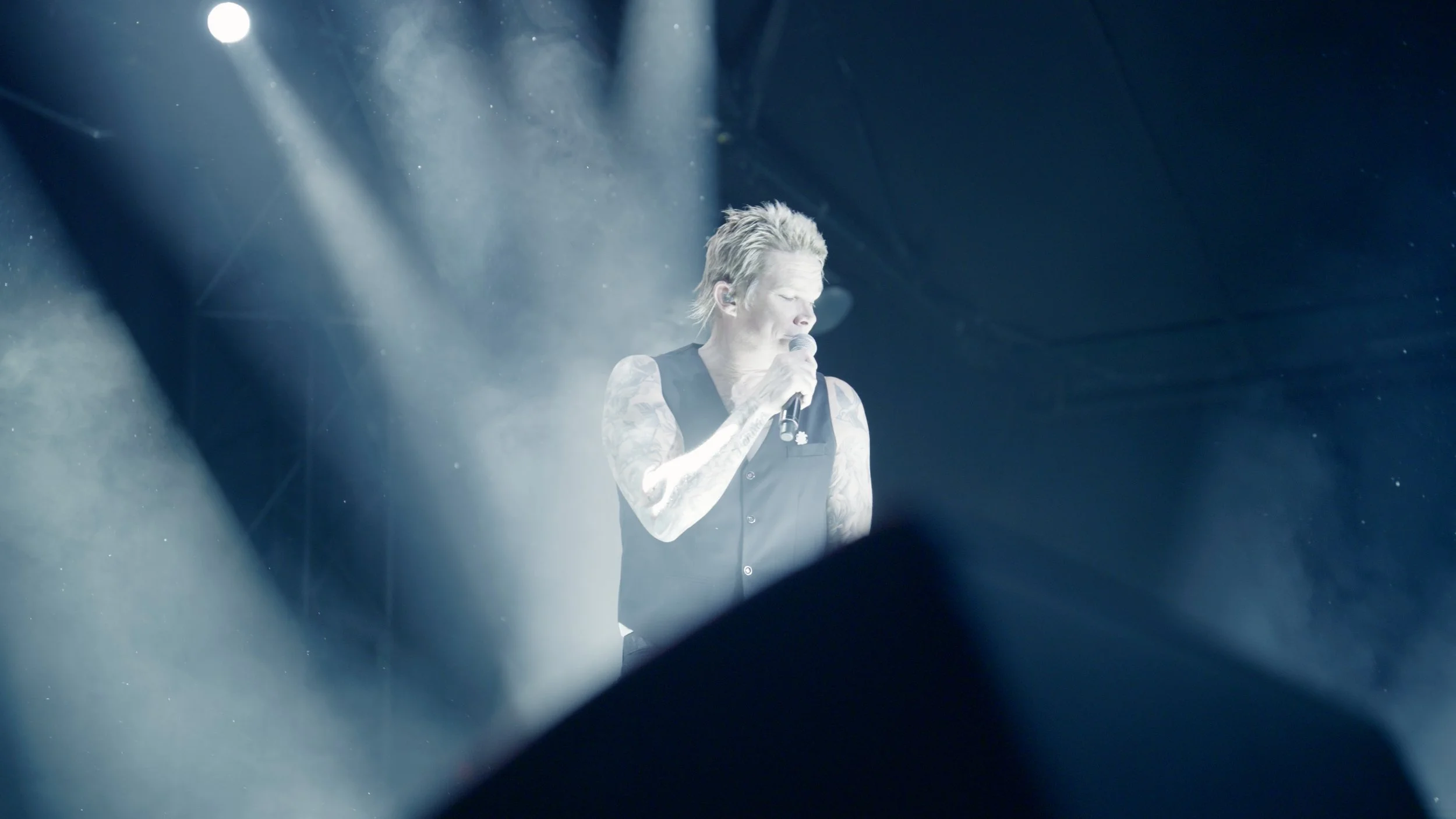 A man with tattoos and spiky blond hair singing into a microphone on stage, with bright spotlights and fog effects.