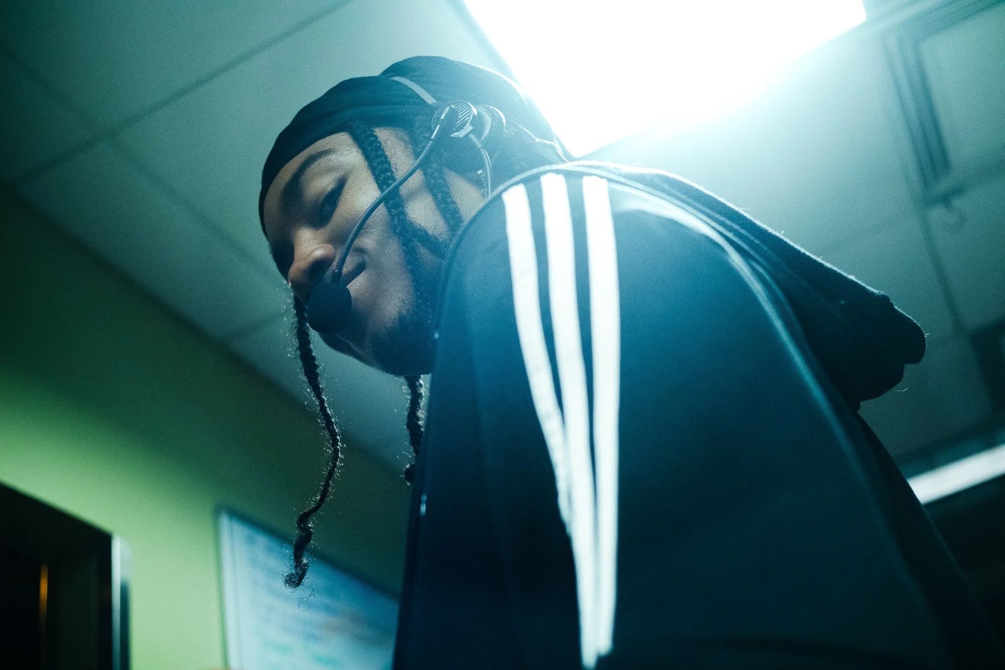 A person with dreadlocks wearing glasses and a dark jacket, looking downwards in a room with bright lighting.