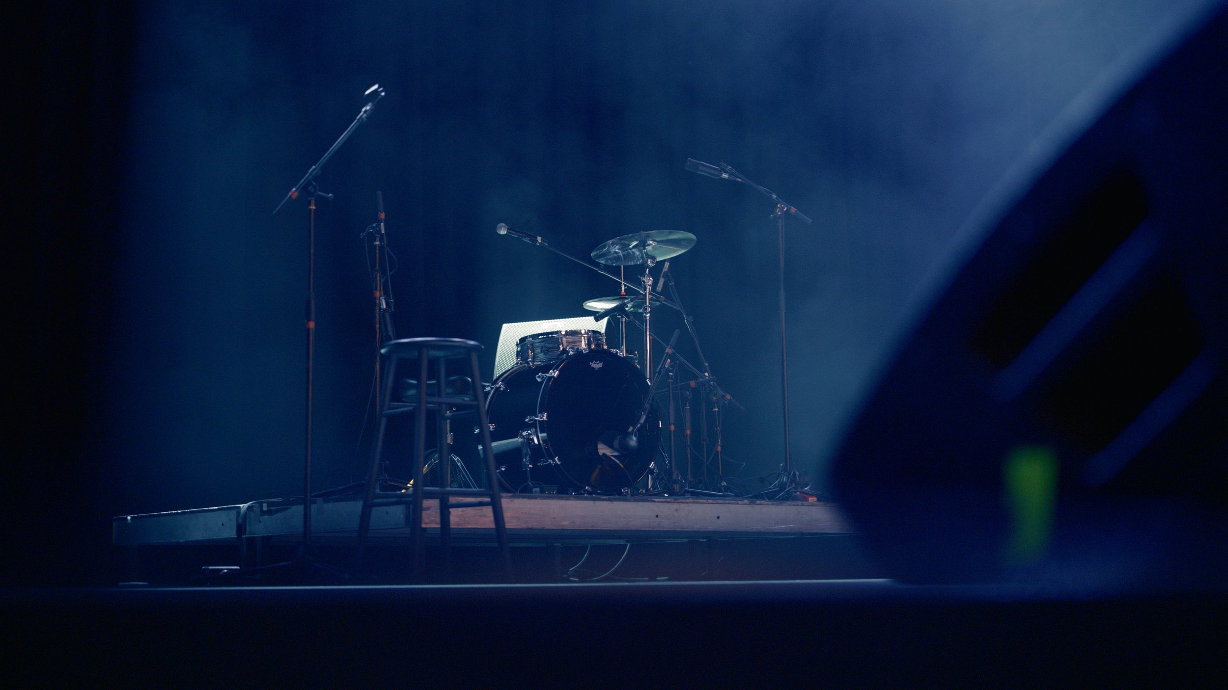 Empty drum set with microphones on stands on stage, illuminated by a spotlight in a dark venue.