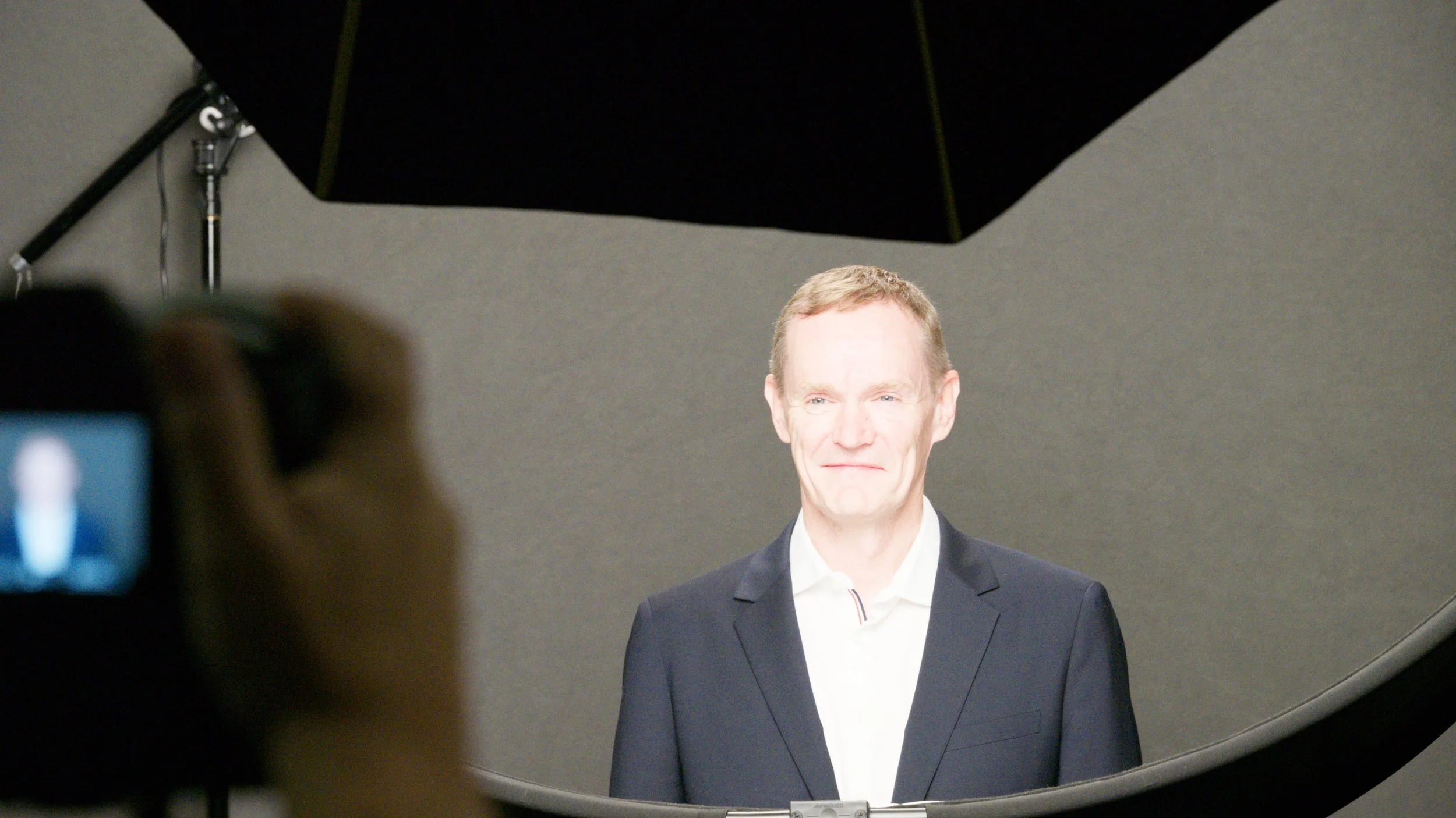 A man in a suit standing in front of a gray background during a photo shoot setup, with studio lighting and a reflector overhead.