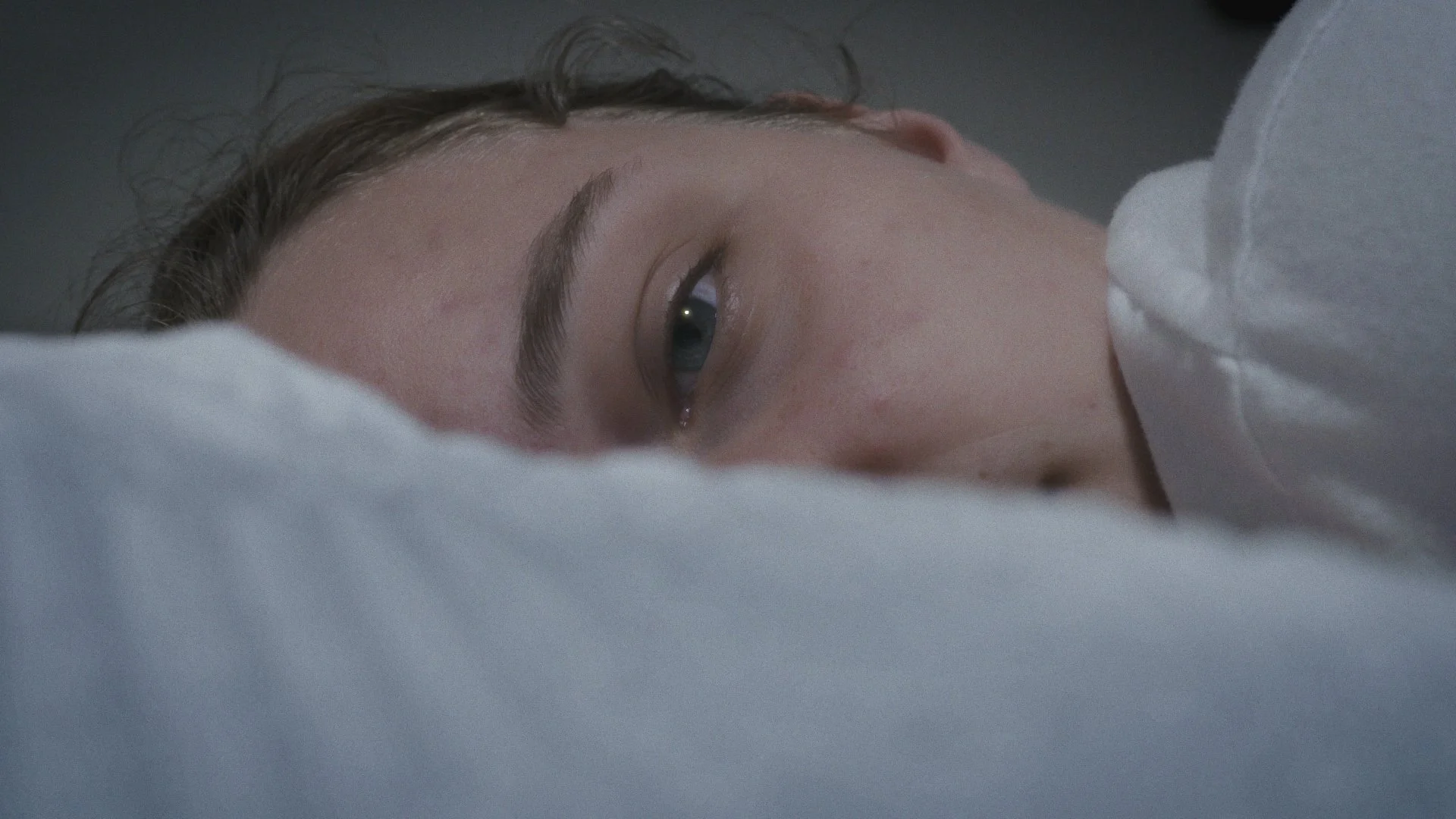 A close-up of a person lying down, partially covered with a white sheet, with only their face visible, showing closed eyes and relaxed facial expression.