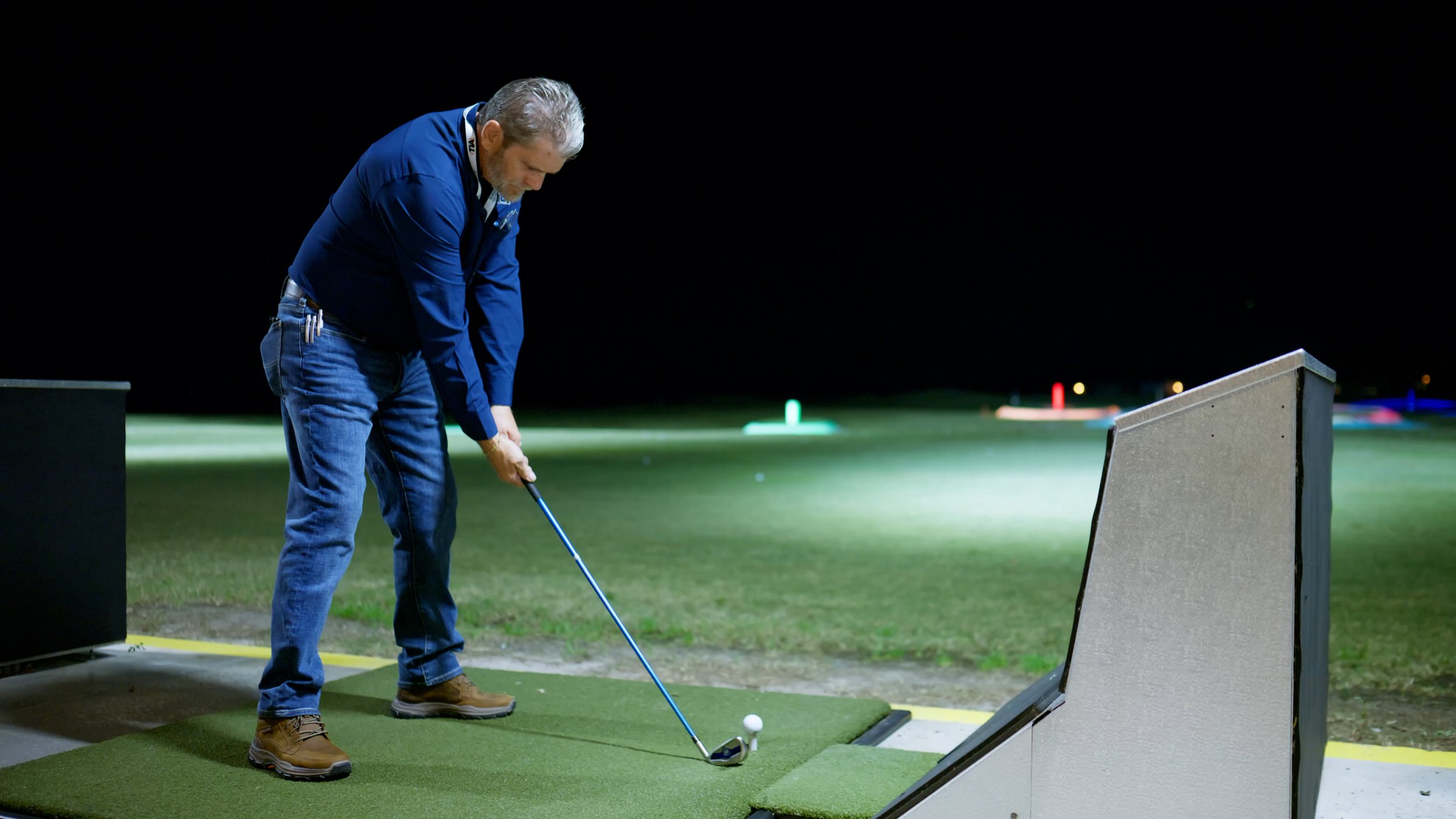 A man in a blue shirt and jeans playing mini golf at night, preparing to hit a golf ball with a putter.