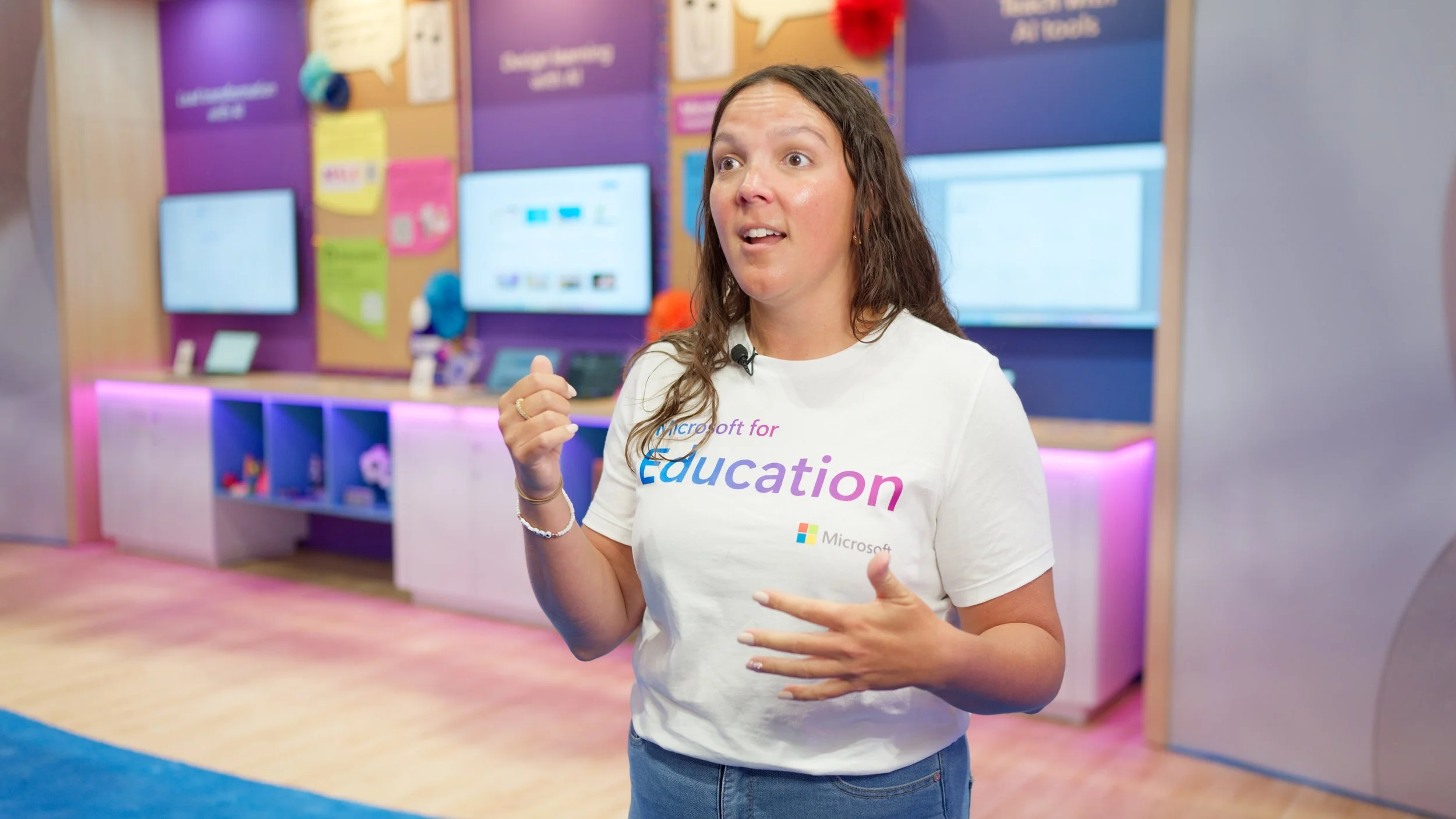 Woman speaking in a classroom or conference setting, wearing a white Microsoft Education T-shirt, with a colorful classroom background and digital screens behind her.
