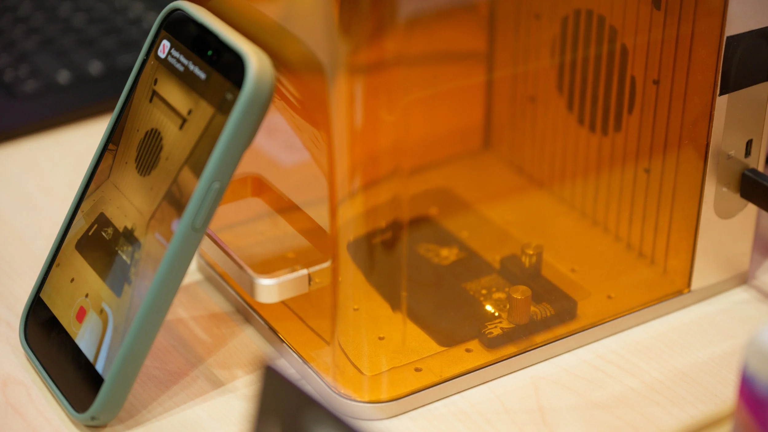 An iPhone positioned to show the screen recording a 3D printer in operation inside a transparent orange enclosure.