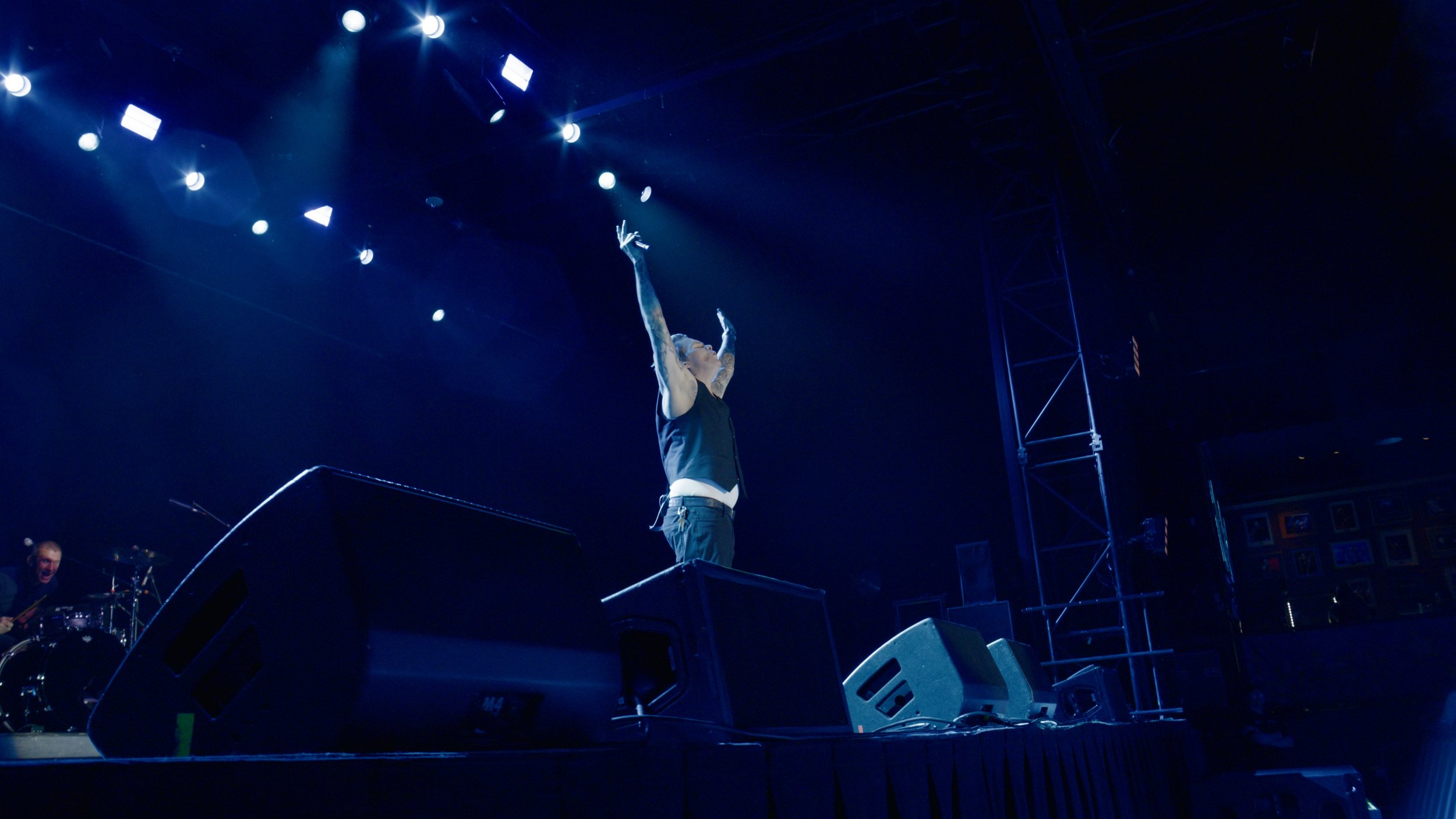 A singer with tattoos on arms performing on stage with arms raised, under bright stage lights, with a drummer visible on the left side of the stage.
