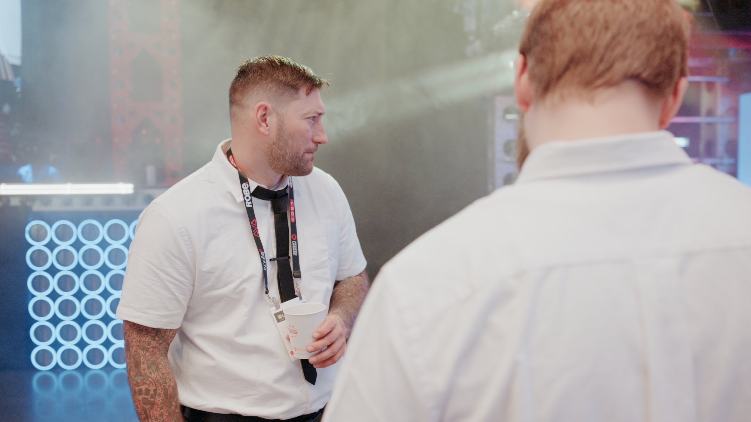 Two men having a conversation at an event. One man is facing the camera, wearing a white shirt and holding a cup. The other man is seen from behind, with red hair and a white shirt.