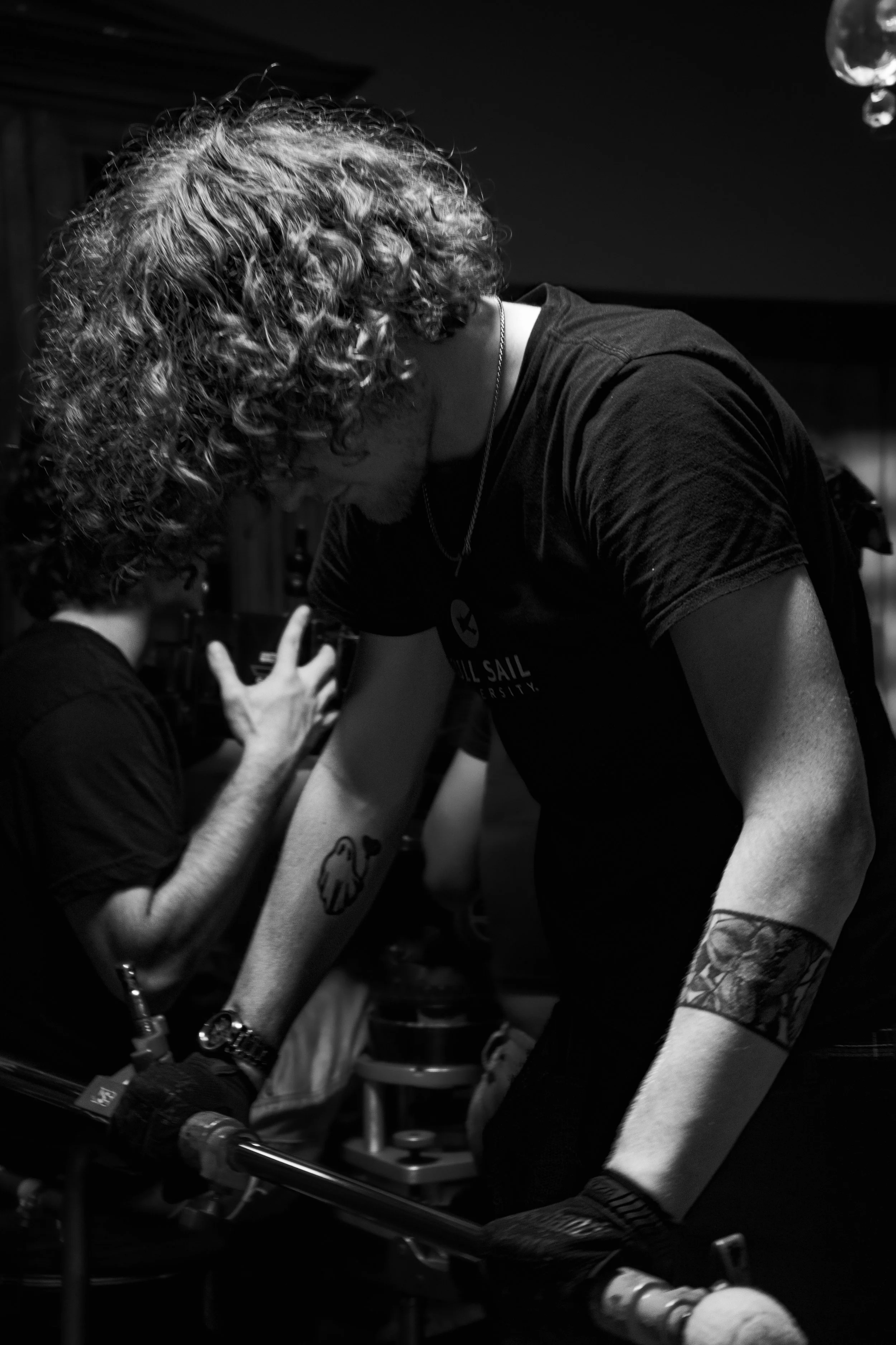 A person with curly hair leaning over a bicycle, with another person in the background engaging in conversation.