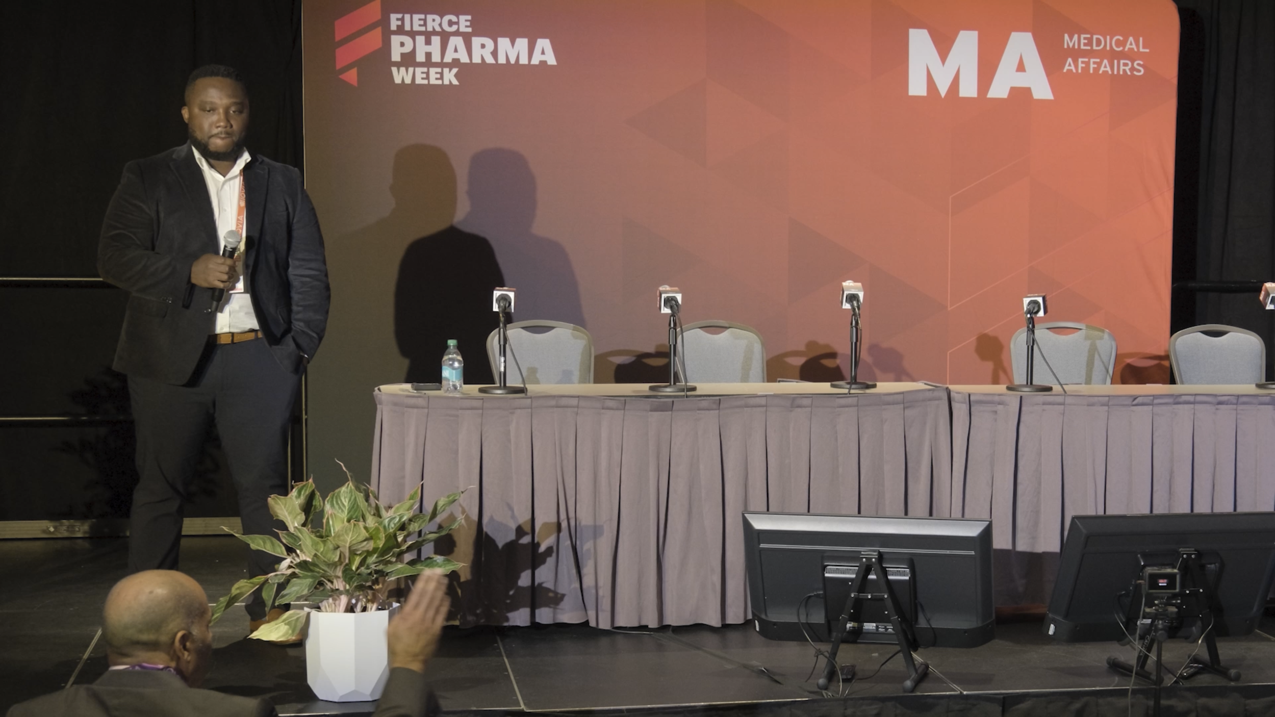A man in a black blazer and white shirt holding a microphone standing next to a table with four microphones, a water bottle, and chairs, at a conference stage with a red backdrop that reads 'Fierce Pharma Week' and 'MA Medical Affairs'.