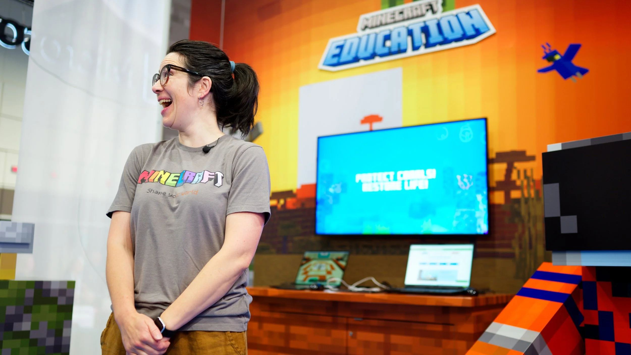 A woman wearing glasses and a gray t-shirt with a colorful logo stands smiling at a Minecraft Education event set up.