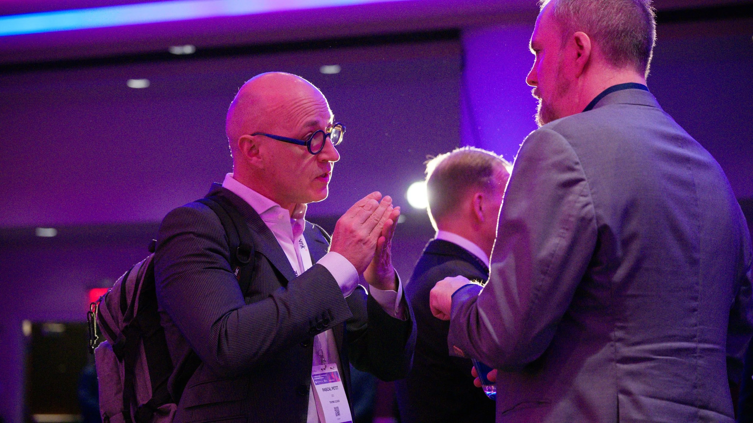 Three men in suits at a conference or event, engaged in conversation and greeting each other with a handshake and prayer-like gesture. Background is dimly lit with purple and blue lighting.
