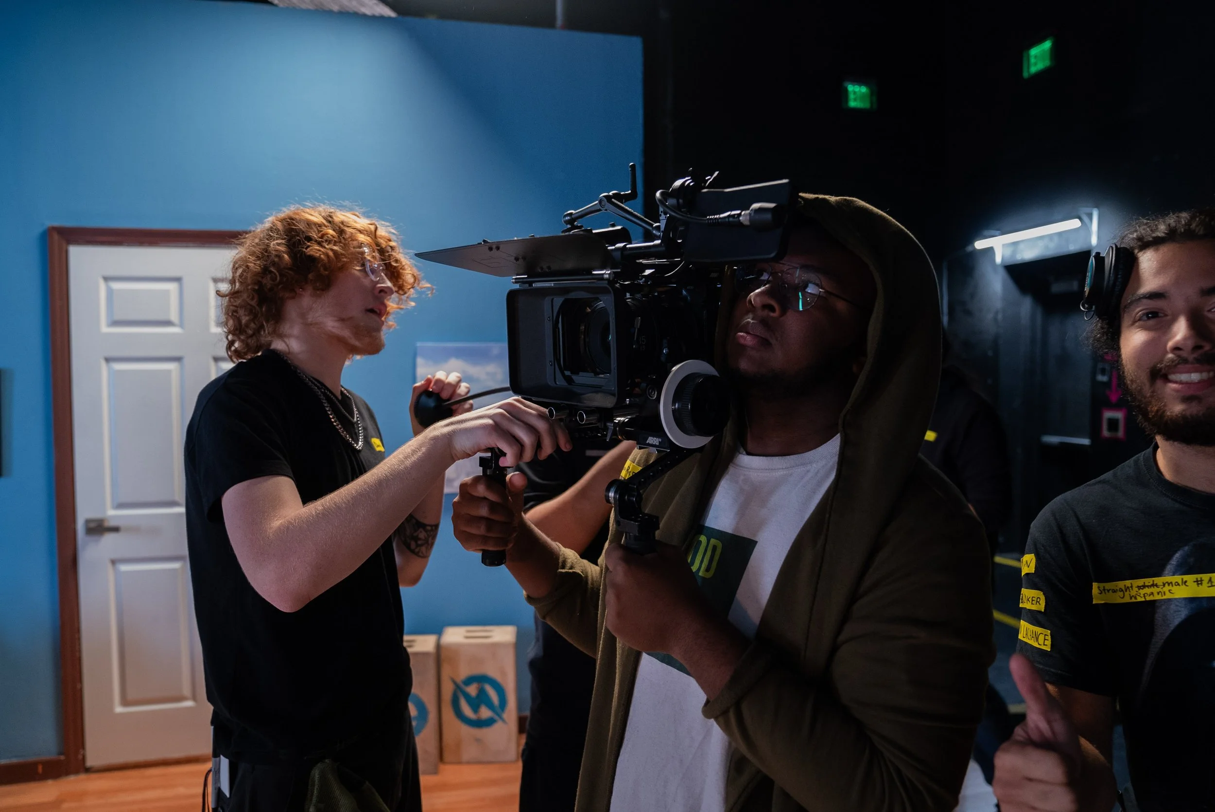 People filming a scene with a professional camera indoors, blue wall, brown door, and cardboard boxes in the background.