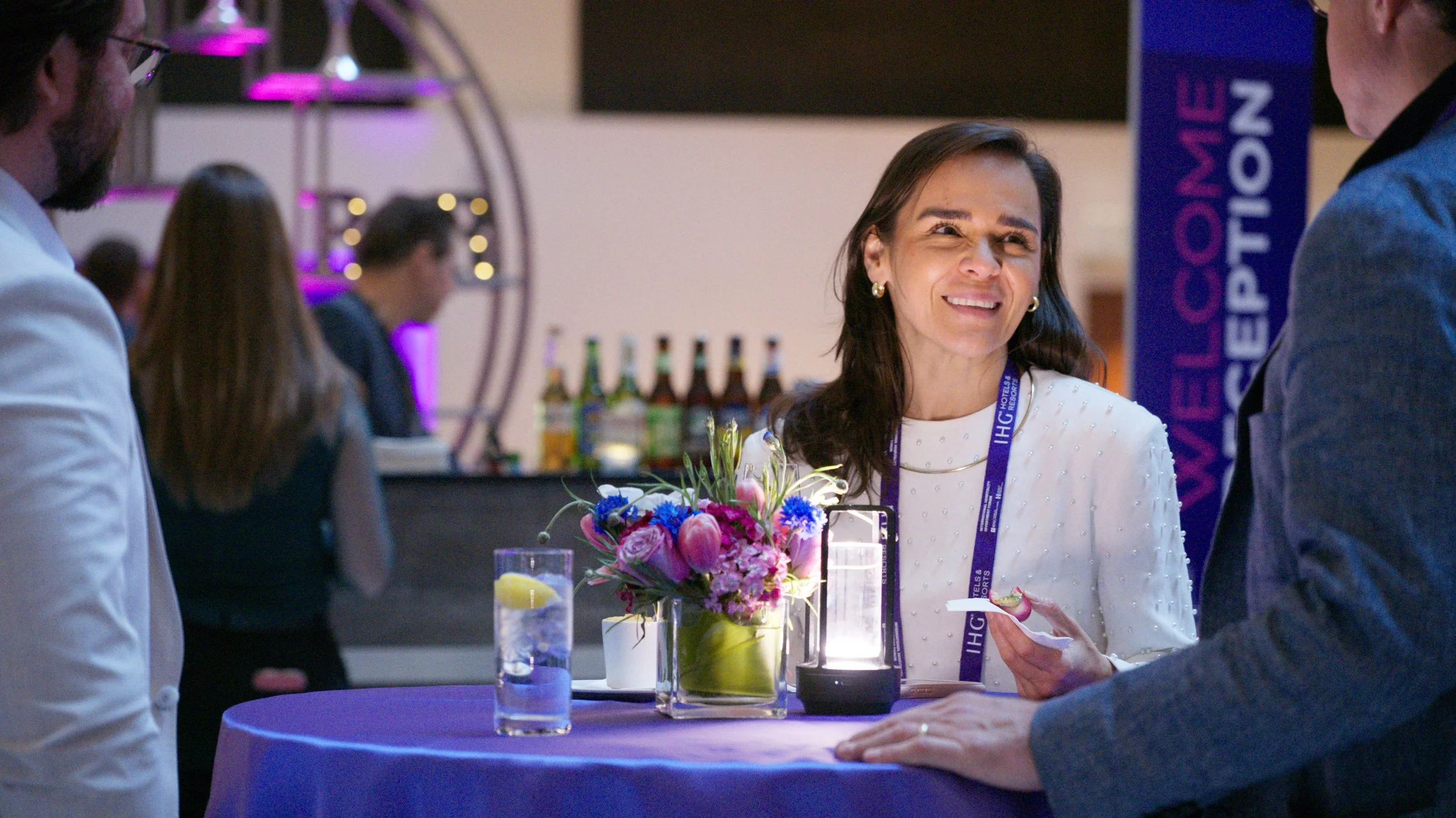 A woman with dark hair and earrings smiling and talking to a man in a formal event, with a bouquet of flowers and drinks on a purple table nearby, and other people and a bar in the background.