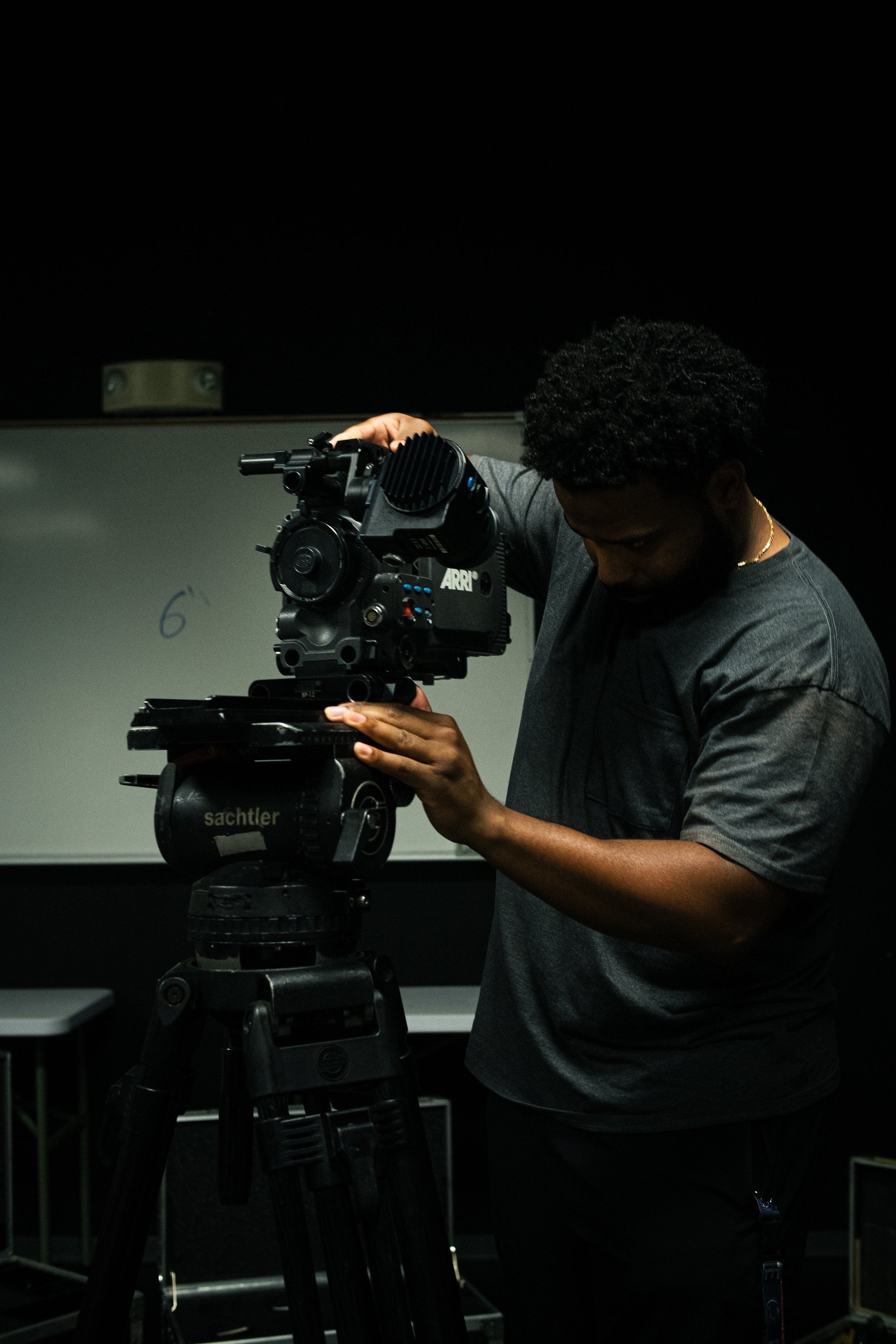 A man operating a professional video camera on a tripod in a dark room, with a whiteboard visible in the background.