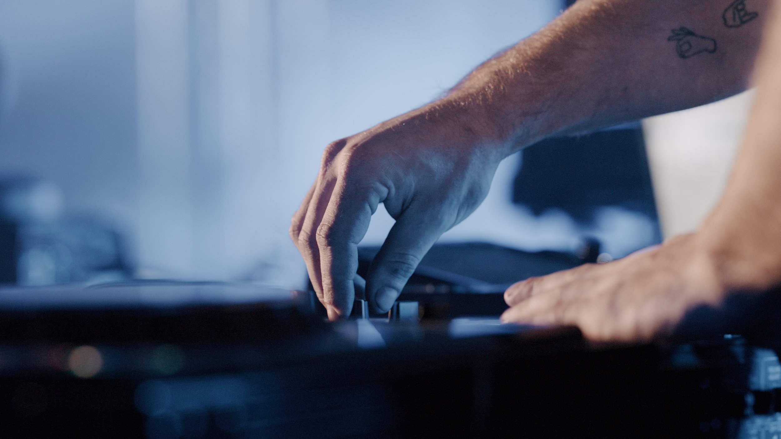 Close-up of a person's hand playing the piano, showing detailed skin texture and tattoos on the arm, with a blurred background.