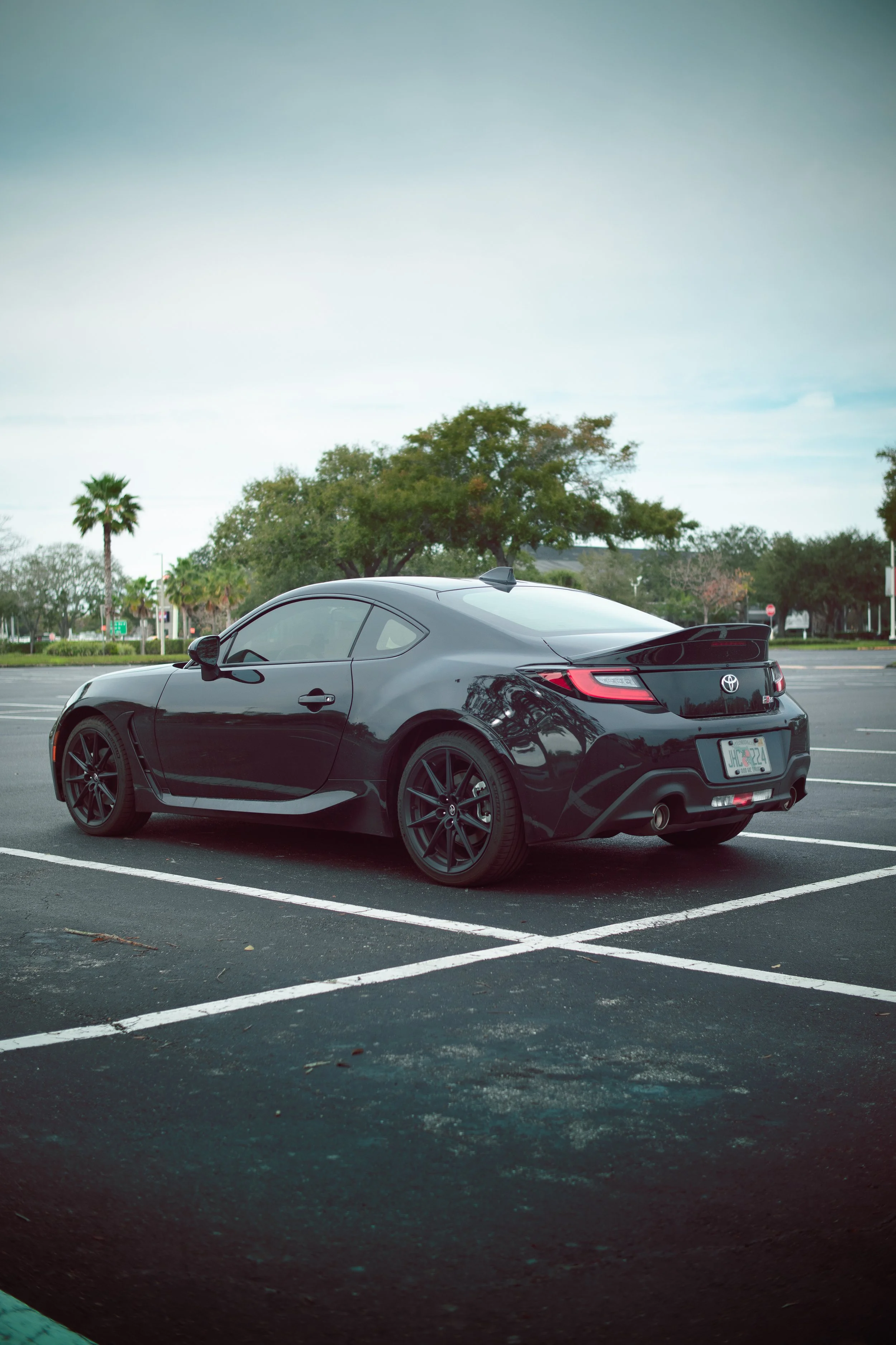 A black Toyota sports car parked in an empty parking lot with trees and a cloudy sky in the background.
