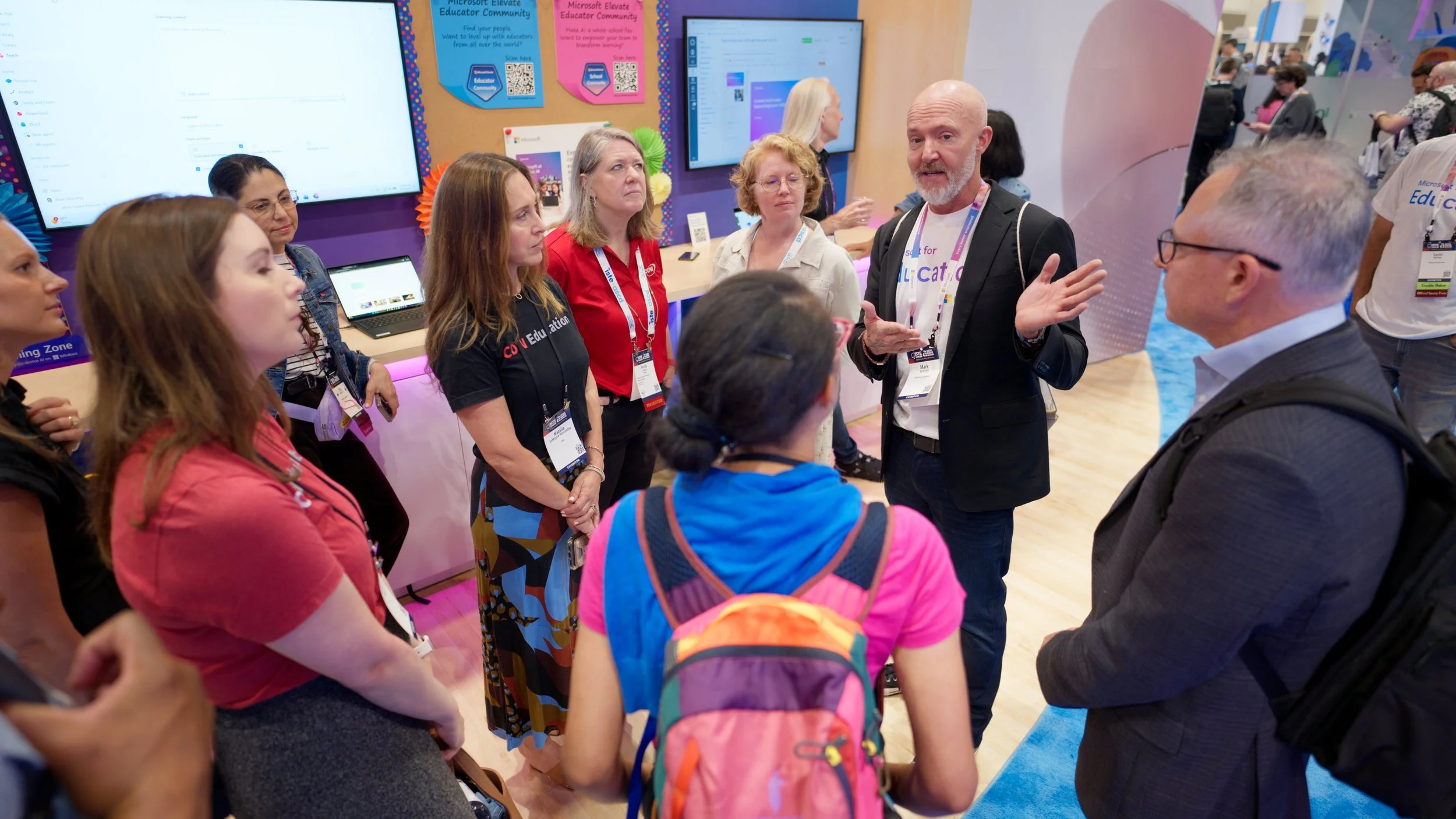 A group of people gathered around a man with a white beard and a black blazer, who is speaking and gesturing with his hands at an educational conference. There are multiple screens and colorful posters in the background.