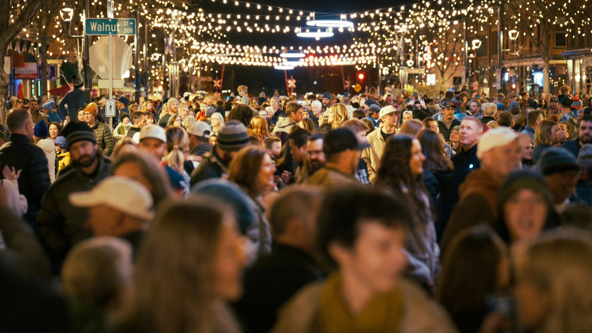 Crowd of people gathered in downtown street decorated with string lights at night.
