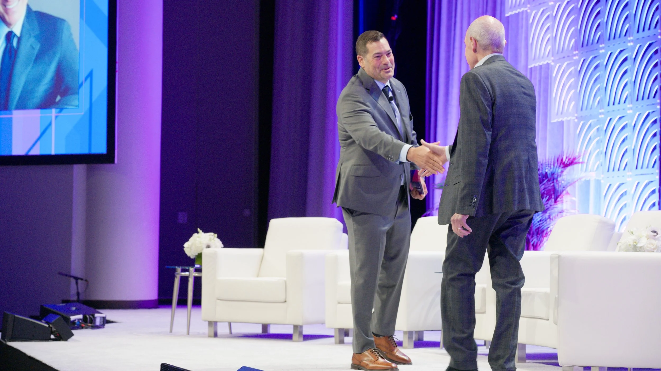 Two men in suits shaking hands on stage at a conference, with a purple and blue backdrop and a large screen to the left showing part of the scene.