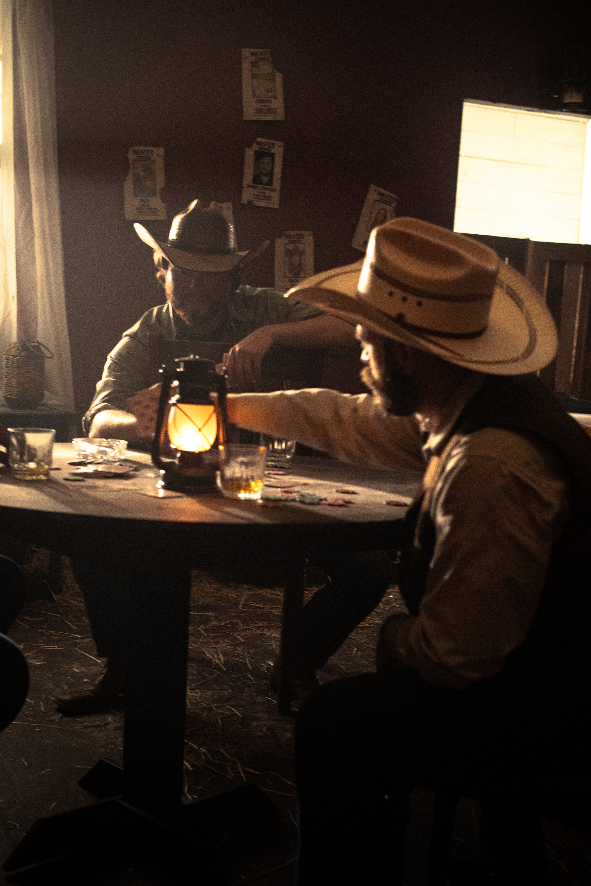 Two men in cowboy hats sitting at a table inside a dimly lit room with a lantern on the table, posters on the wall, and a window with bright light.