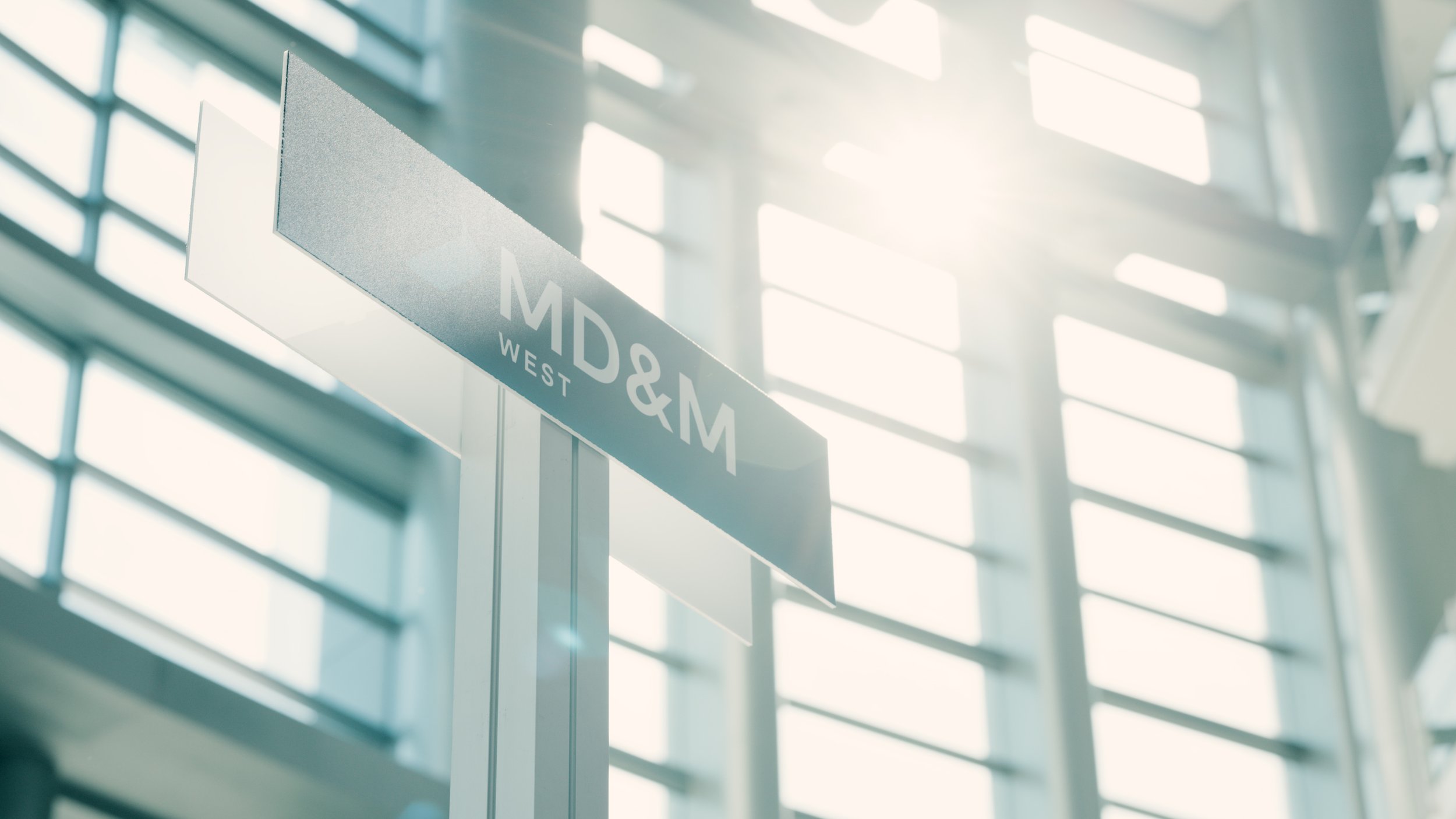 A sign with the text 'MD&M WEST' hanging inside a modern building with large glass windows and sunlight streaming in.