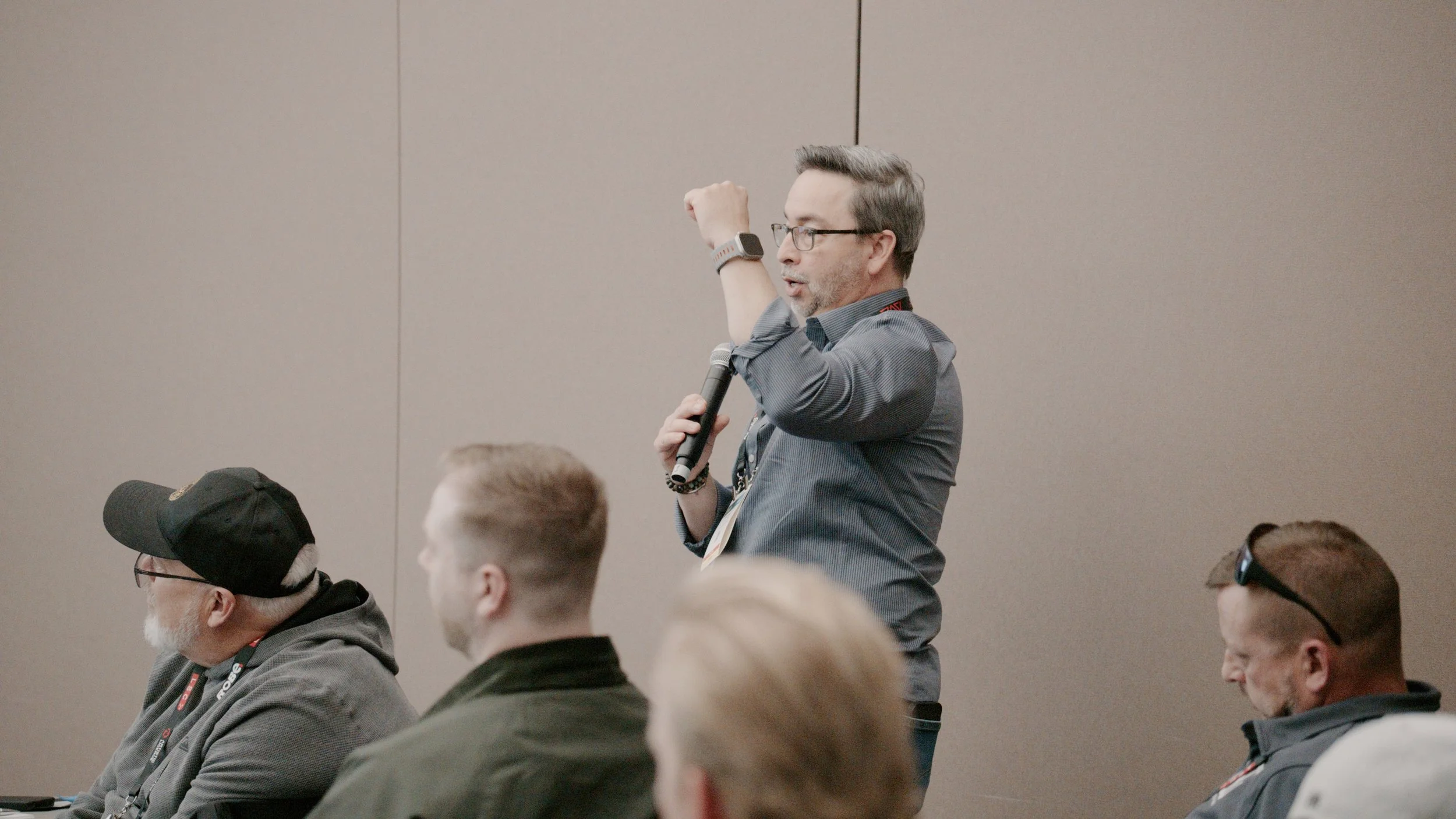 A man wearing glasses and a gray shirt is standing and speaking into a microphone, raising his left hand with a clenched fist, during a conference or meeting, with an audience of seated people in front of him.