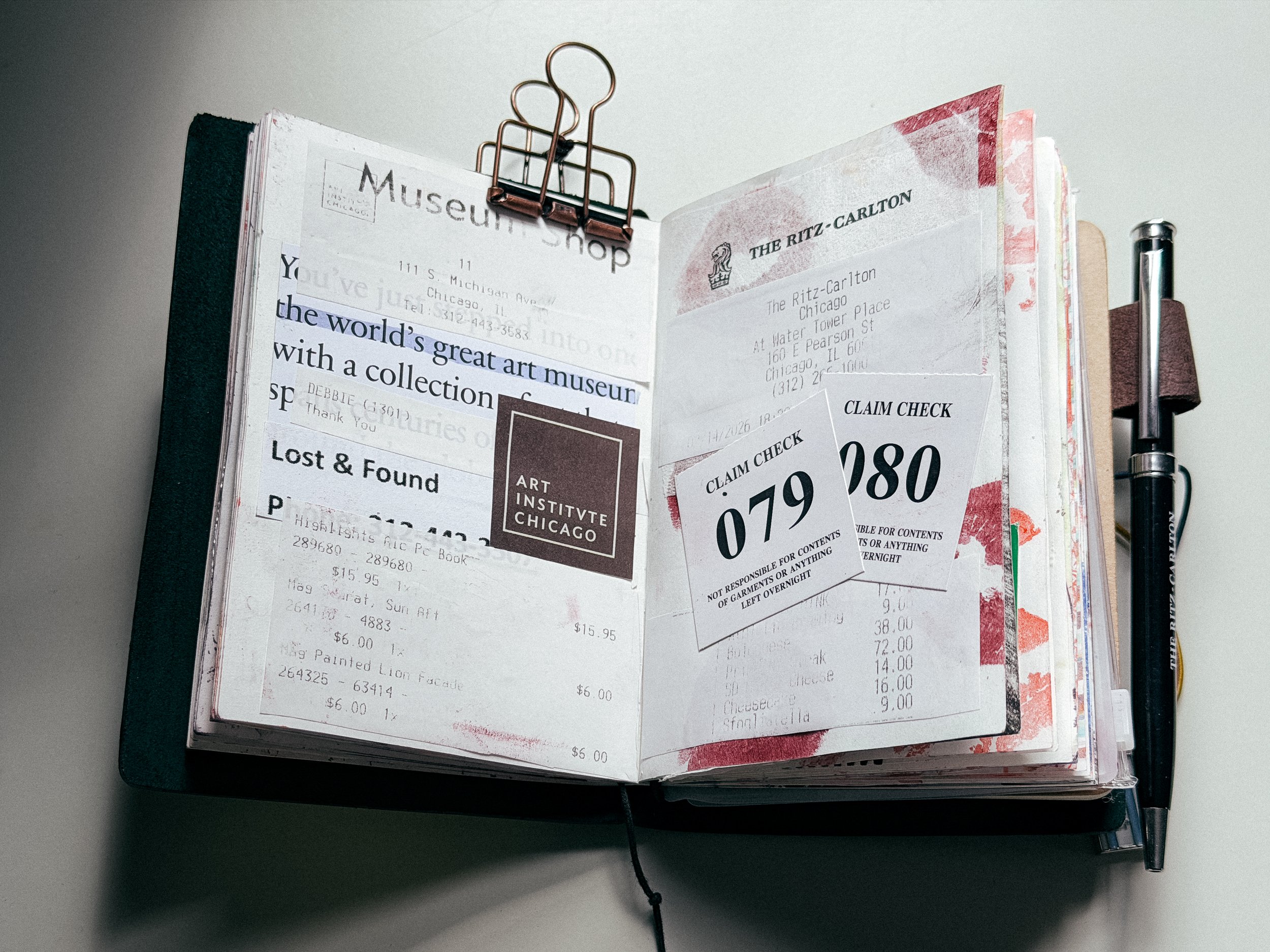 Open scrapbook or journal with various receipts, ticket stubs, and notes attached, held together by a large copper-colored binder clip, and a pen attached on the right side.