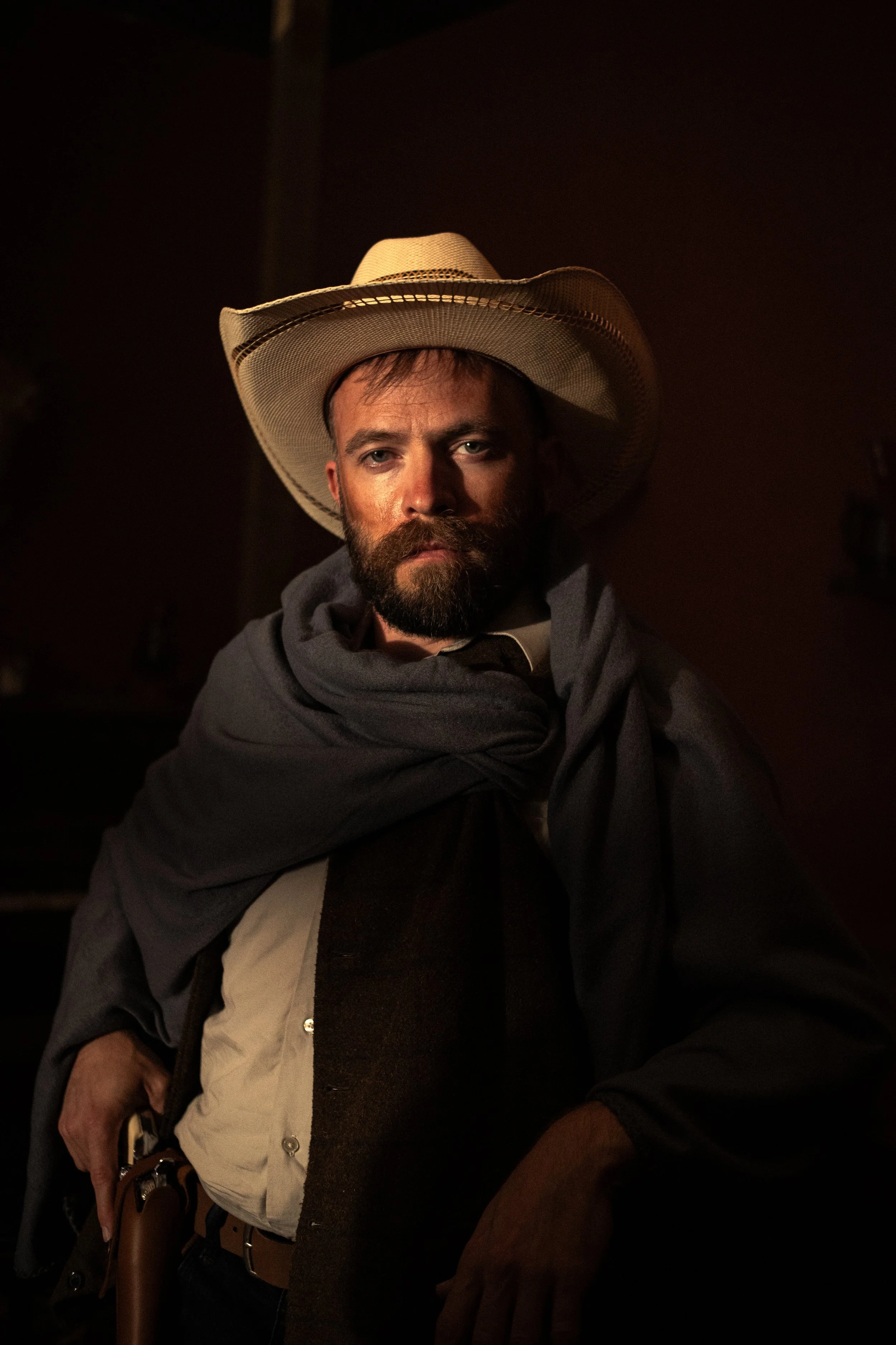 A man with a beard wearing a large straw cowboy hat, a dark hoodie, and a layered shirt, sitting in a dimly lit environment.