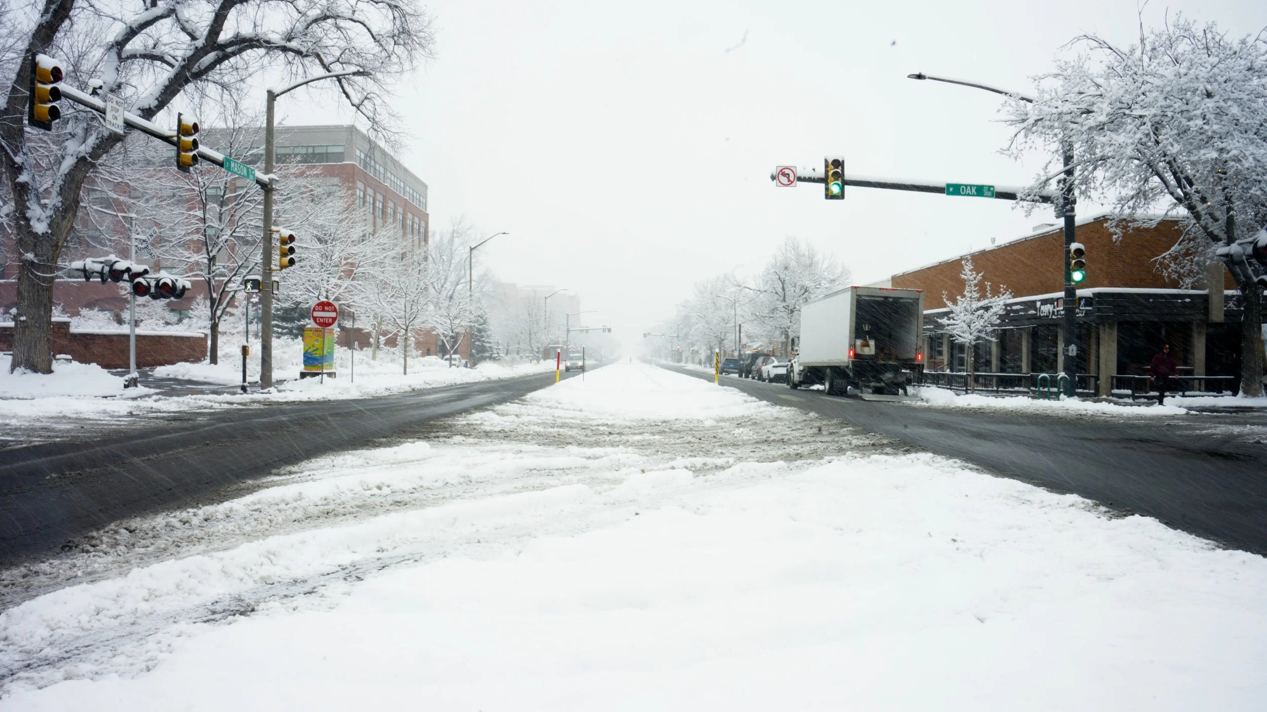 Snow-covered city street with traffic lights, parked cars, trees, and buildings in winter weather.
