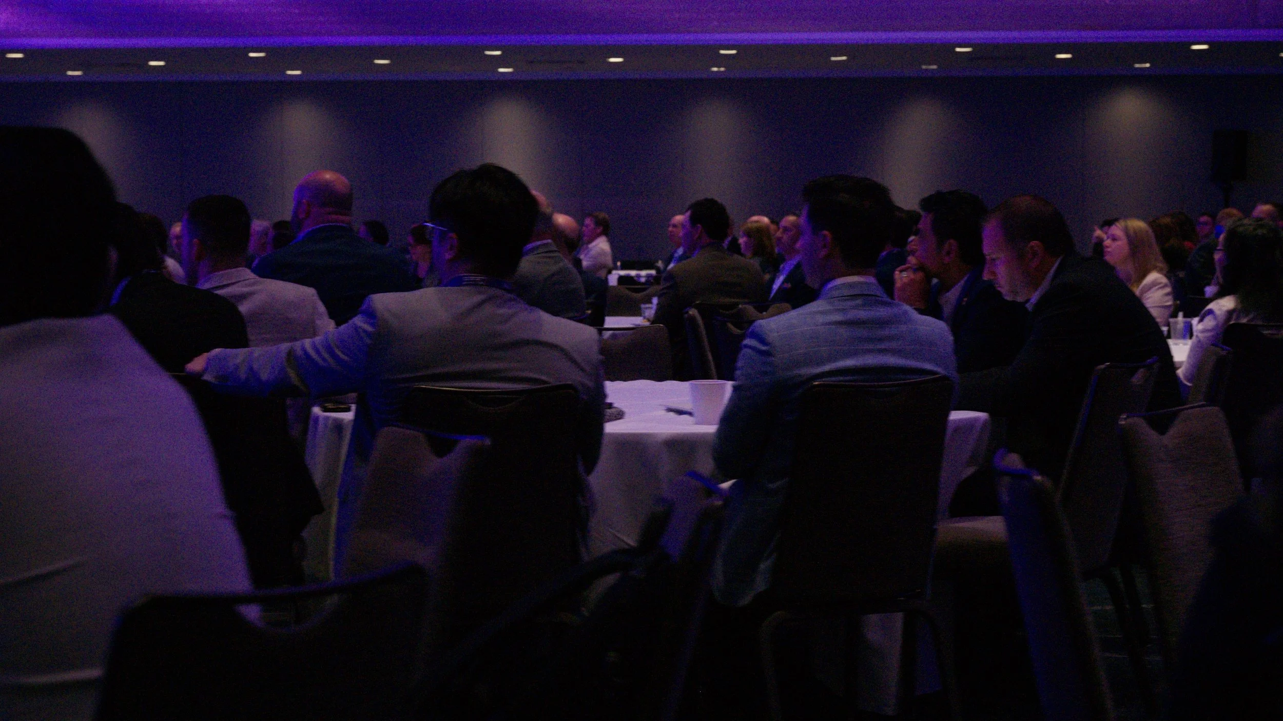 People attending a conference in a dimly lit room, seated at round tables, listening to a presentation.