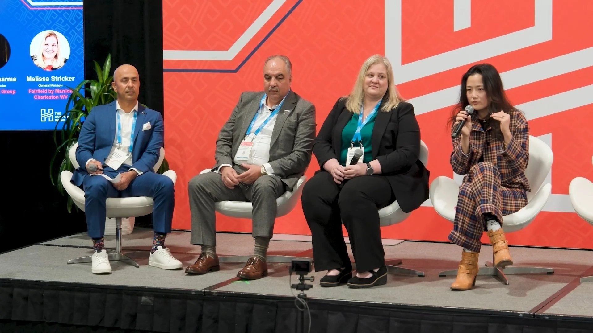 Four people sitting on stage in a panel discussion. The first person on the left is a man in a blue suit and white shoes, holding a microphone. Next is a man in a gray suit with brown shoes. The third is a woman in a black blazer and green top, holdi