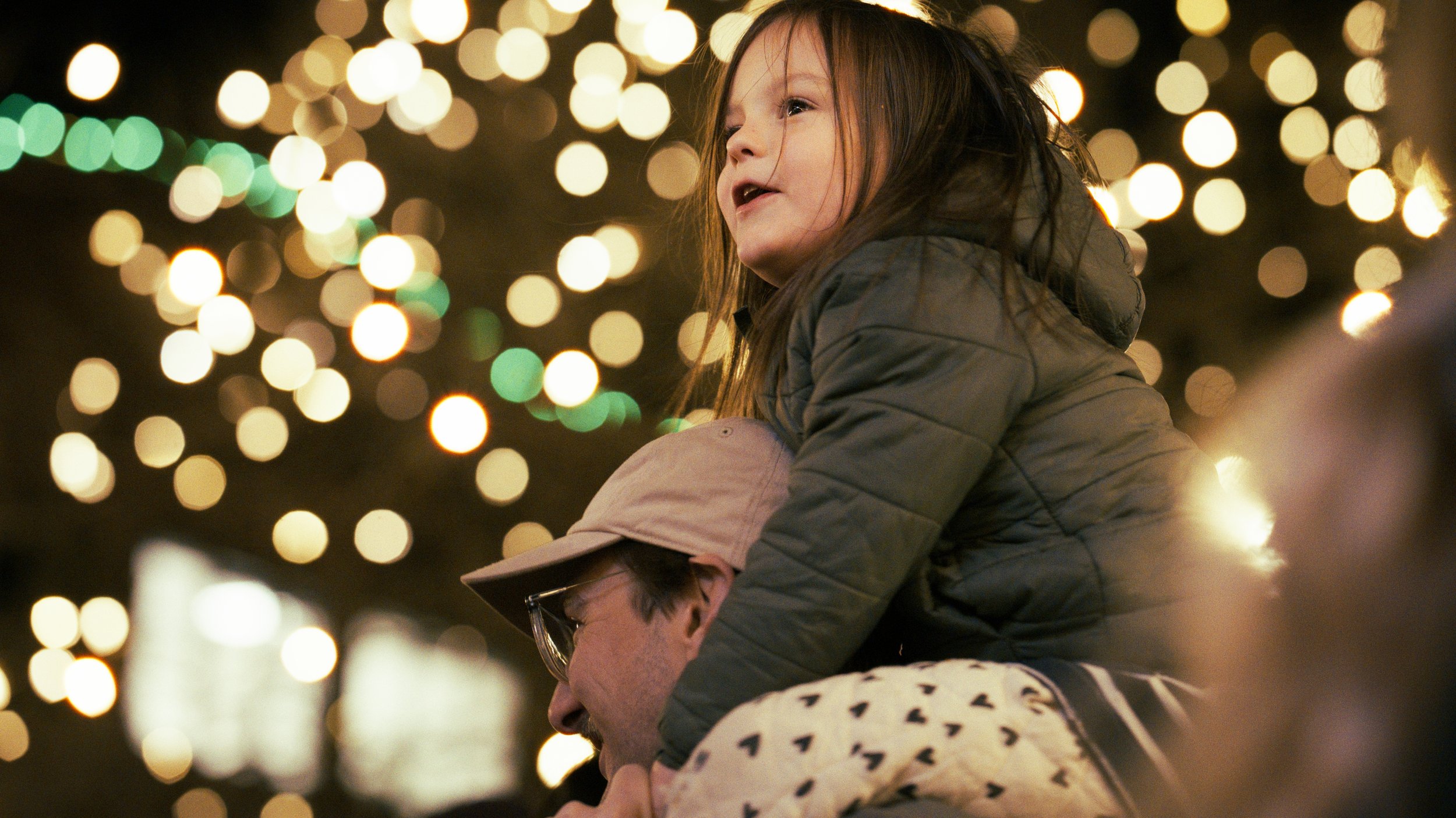 A young girl sitting on an adult's shoulders during nighttime, surrounded by blurred festive lights, with her face turned slightly to the side.