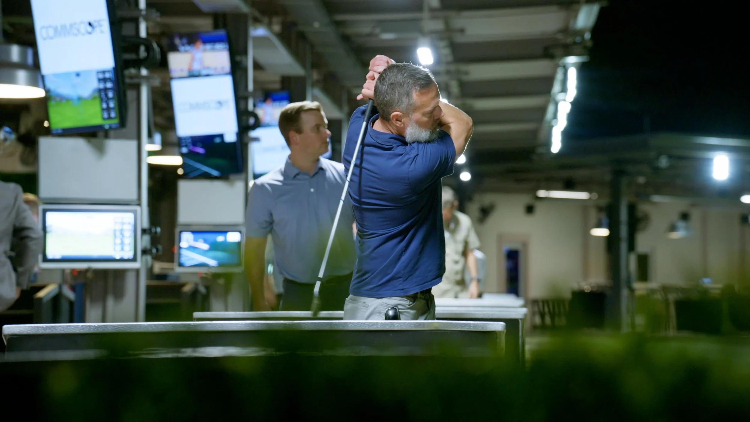 A man with gray hair and beard wearing a navy blue shirt is swinging a golf club in an indoor golf practice facility. There are multiple monitors displaying golf courses and statistics, and a young man in a light blue shirt observing in the backgroun