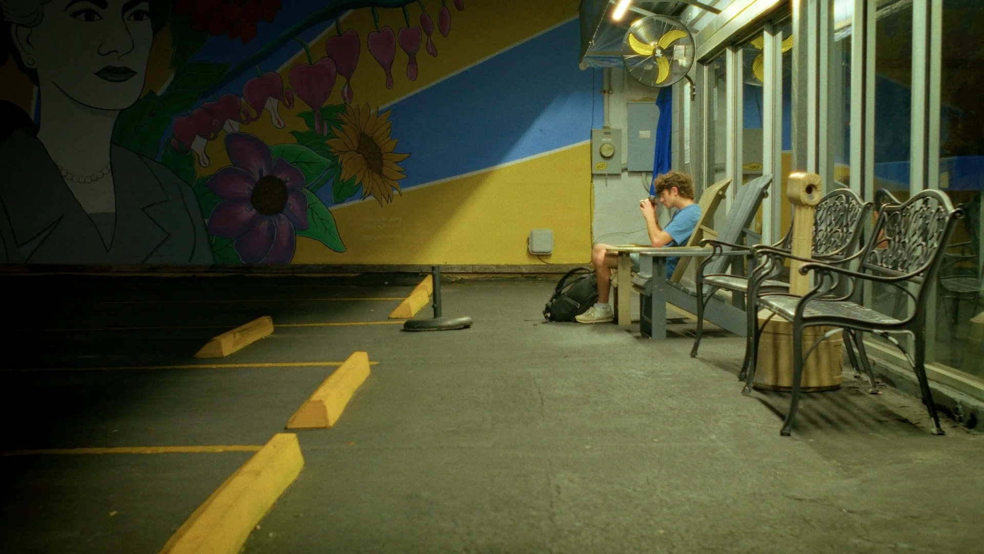A young man in a blue t-shirt and shorts sitting on a bench at a bus stop, looking at his phone, with a backpack on the ground next to him. The background features colorful wall art of a woman's face and flowers, and there are empty metal benches to 