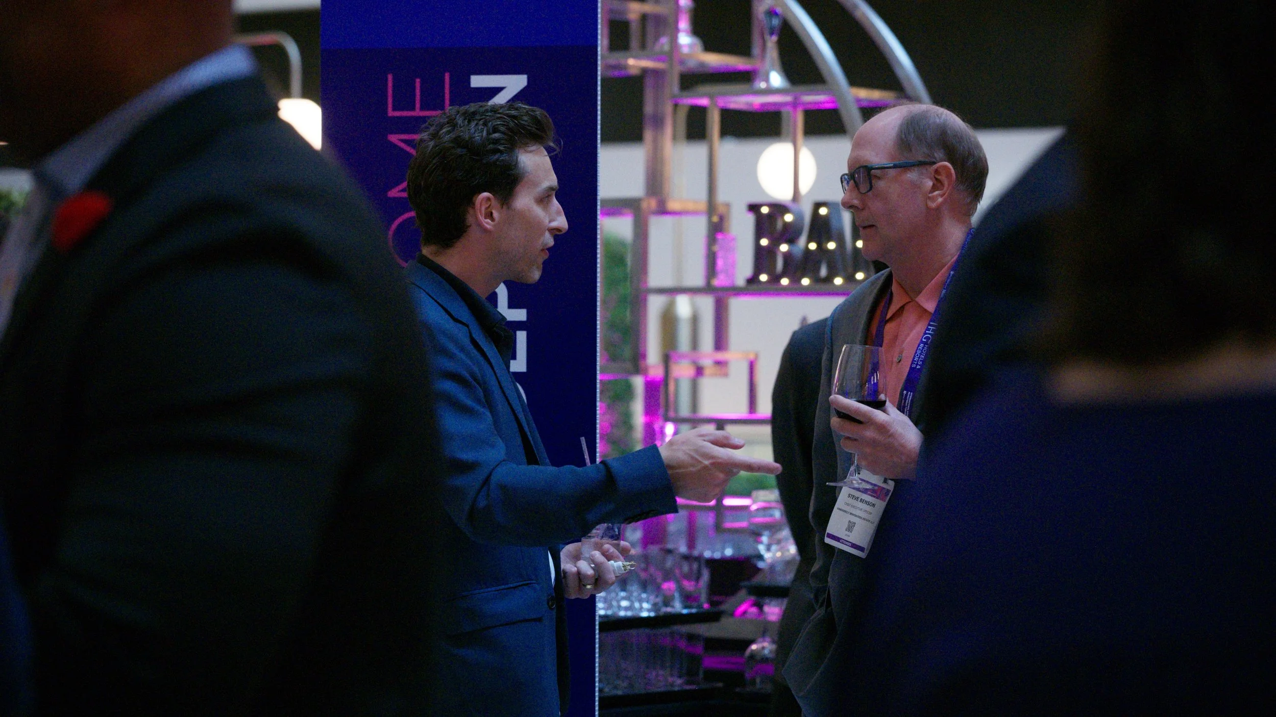 Two men in business attire are engaged in a conversation at an event, with one holding a glass of red wine. They are standing near a display with purple and pink lighting, and there are blurred figures and a bar or display setup in the background.