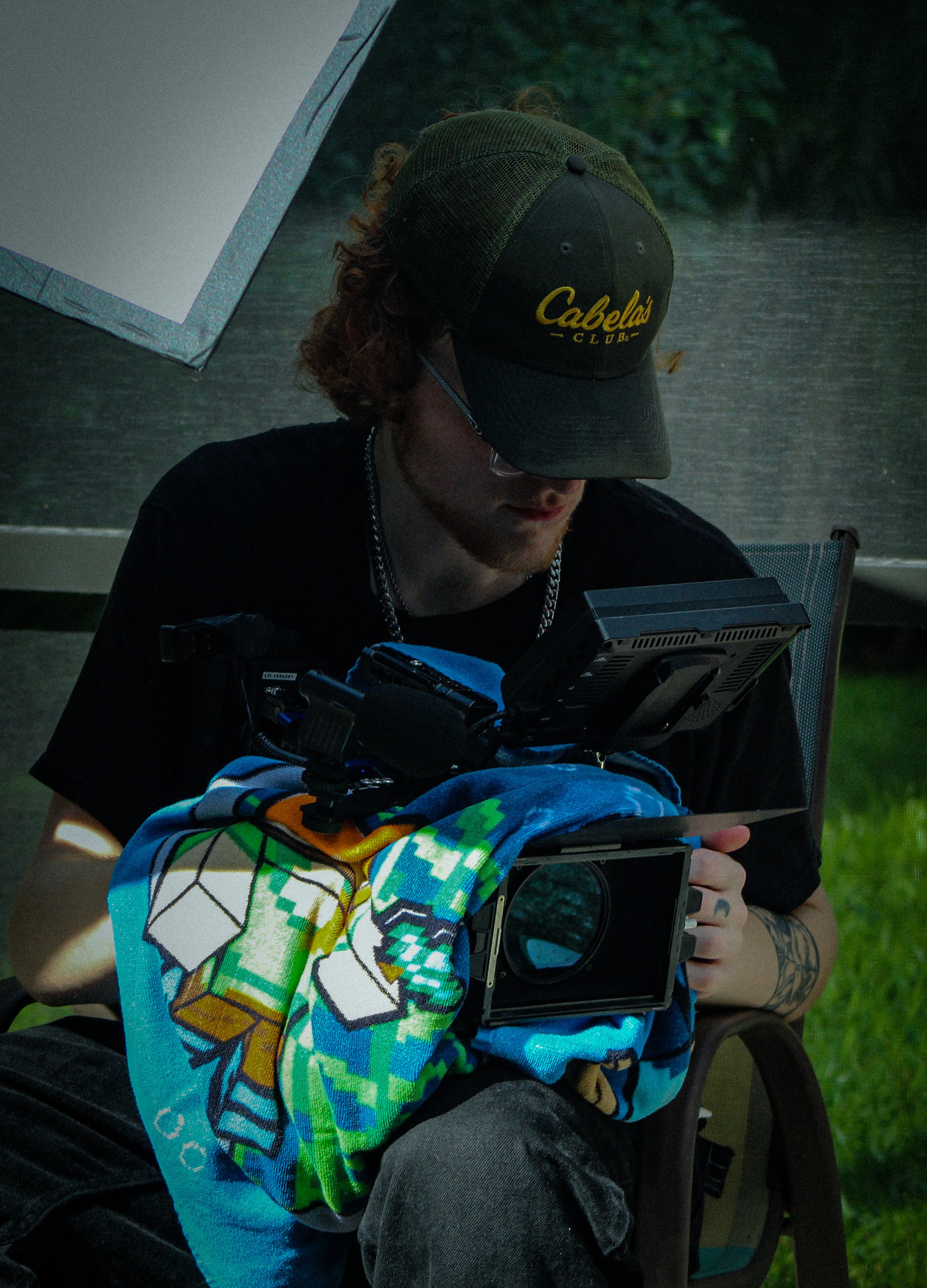 Person with curly hair wearing a Cabela's hat, glasses, and a black shirt, sitting outdoors, holding a camera and a colorful towel.