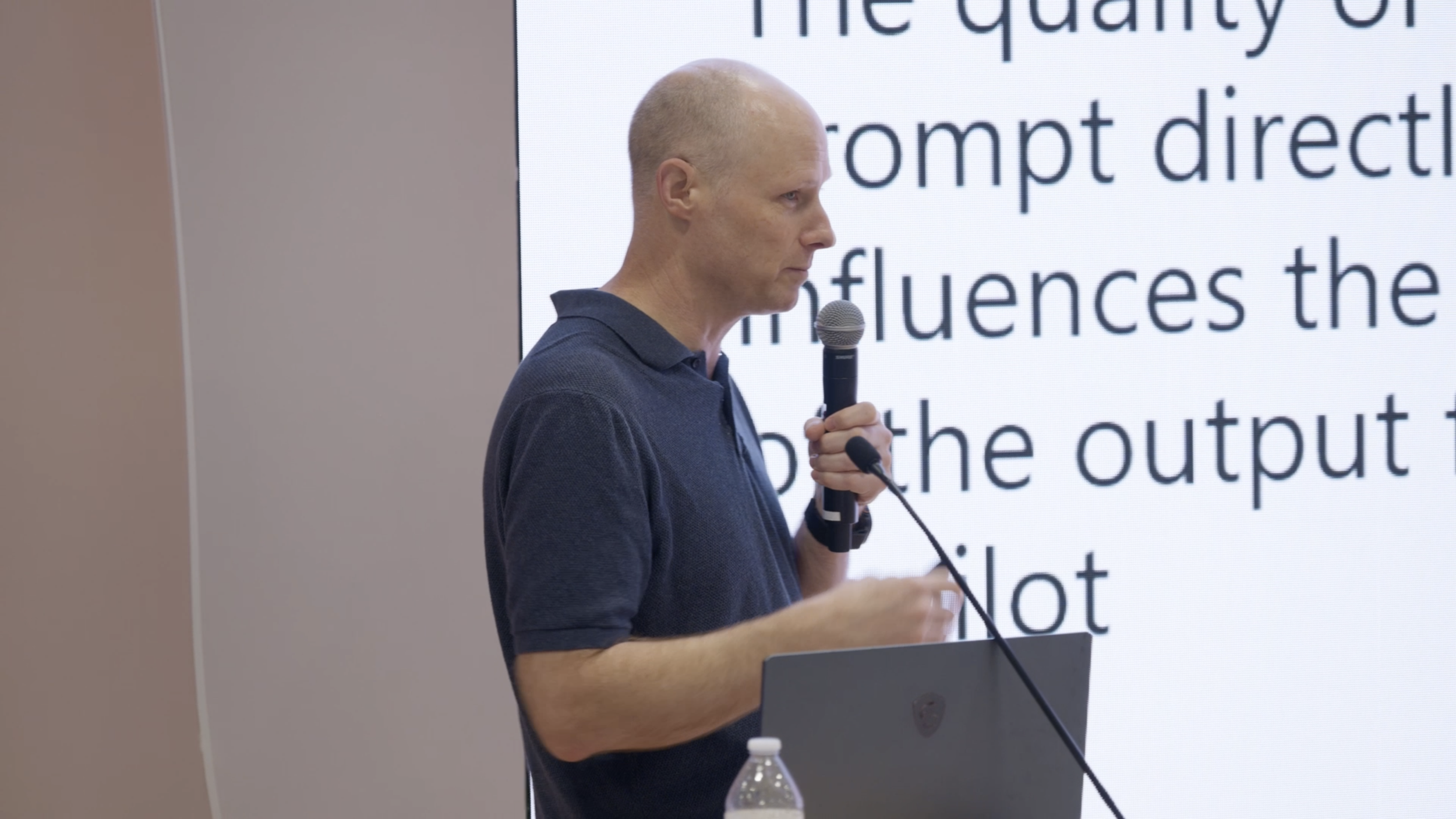 A bald man wearing a navy polo shirt holding a microphone and standing in front of a large screen with text.