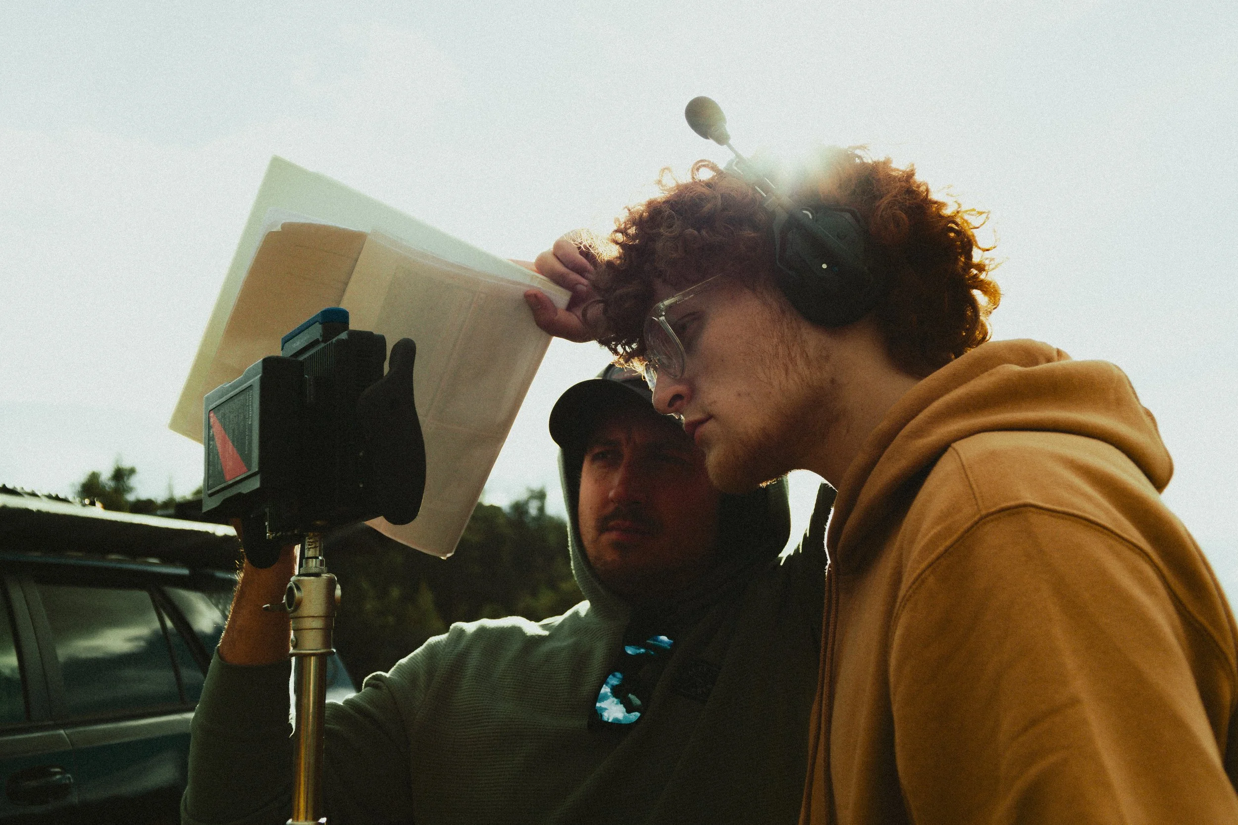 Two men working outdoors with a camera mounted on a tripod, one adjusting the camera while the other observes, during daylight.