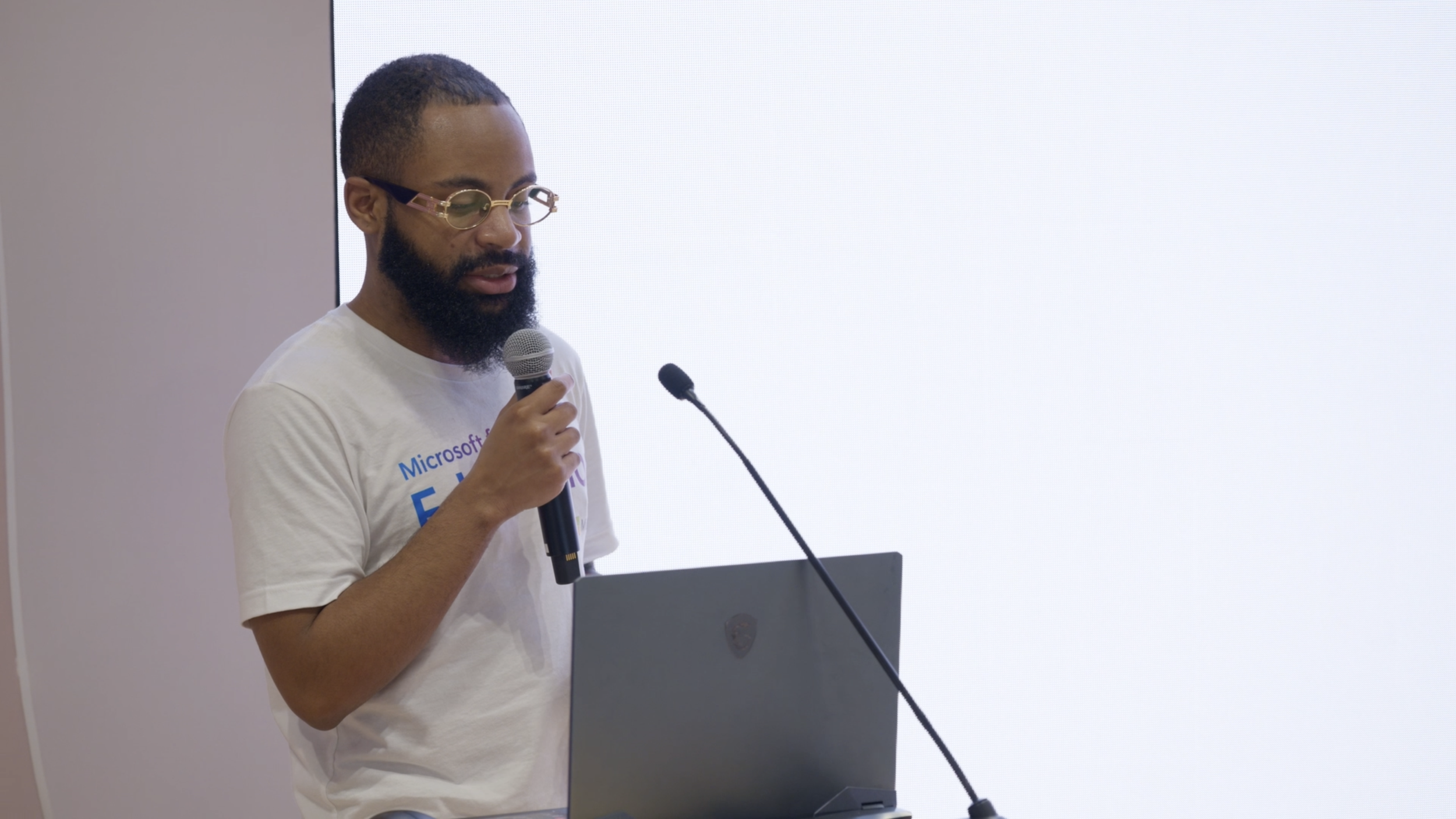 A man with glasses and a beard is speaking into a microphone at a podium during a presentation. He wears a white T-shirt with some text on it and stands in front of a large, white, blank screen.