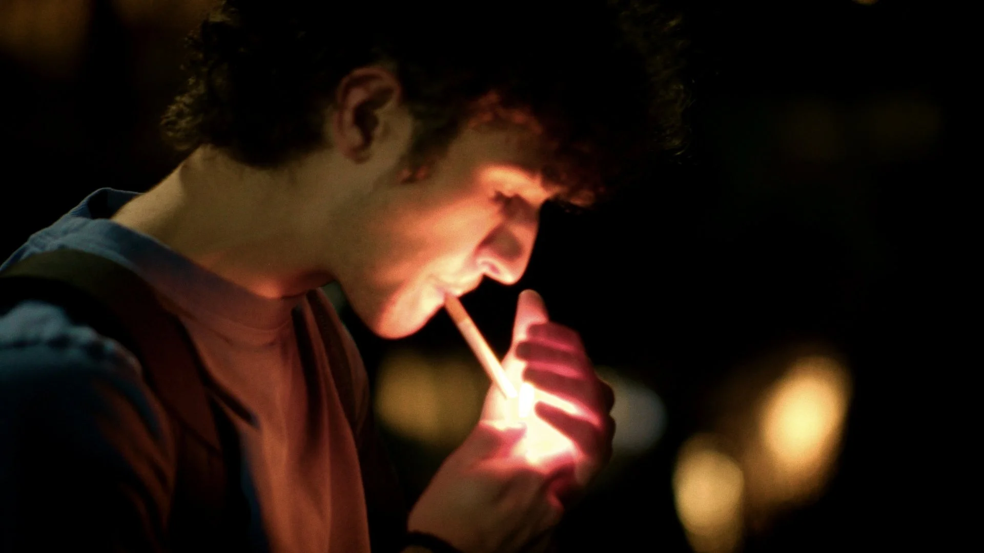 A young man with curly hair lighting a cigarette at night, with blurred city lights in the background.