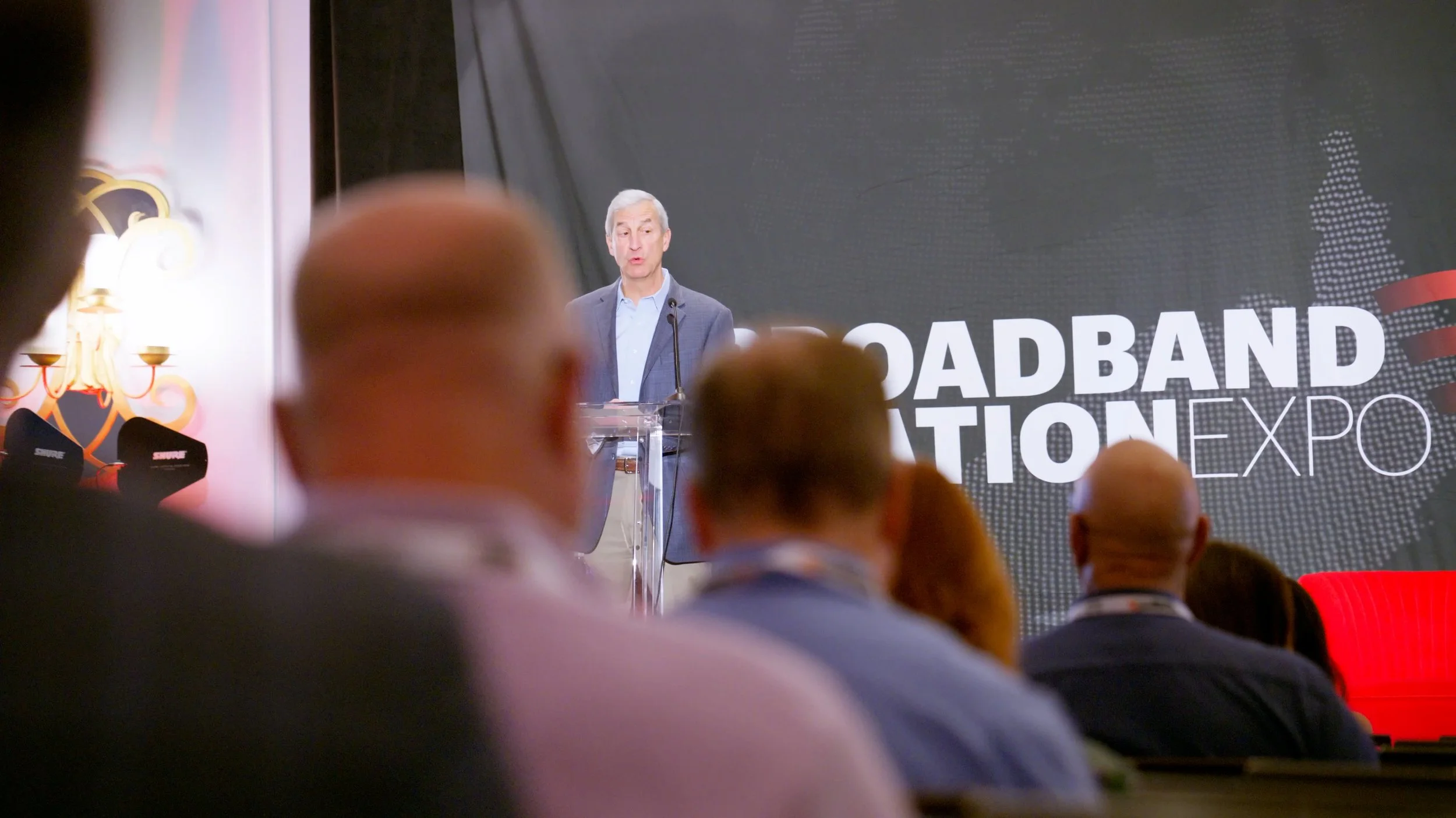 A man in a suit speaking at a podium during a keynote address at the Roadband Automation Expo, with an audience seated in front of him and a large event banner behind him.