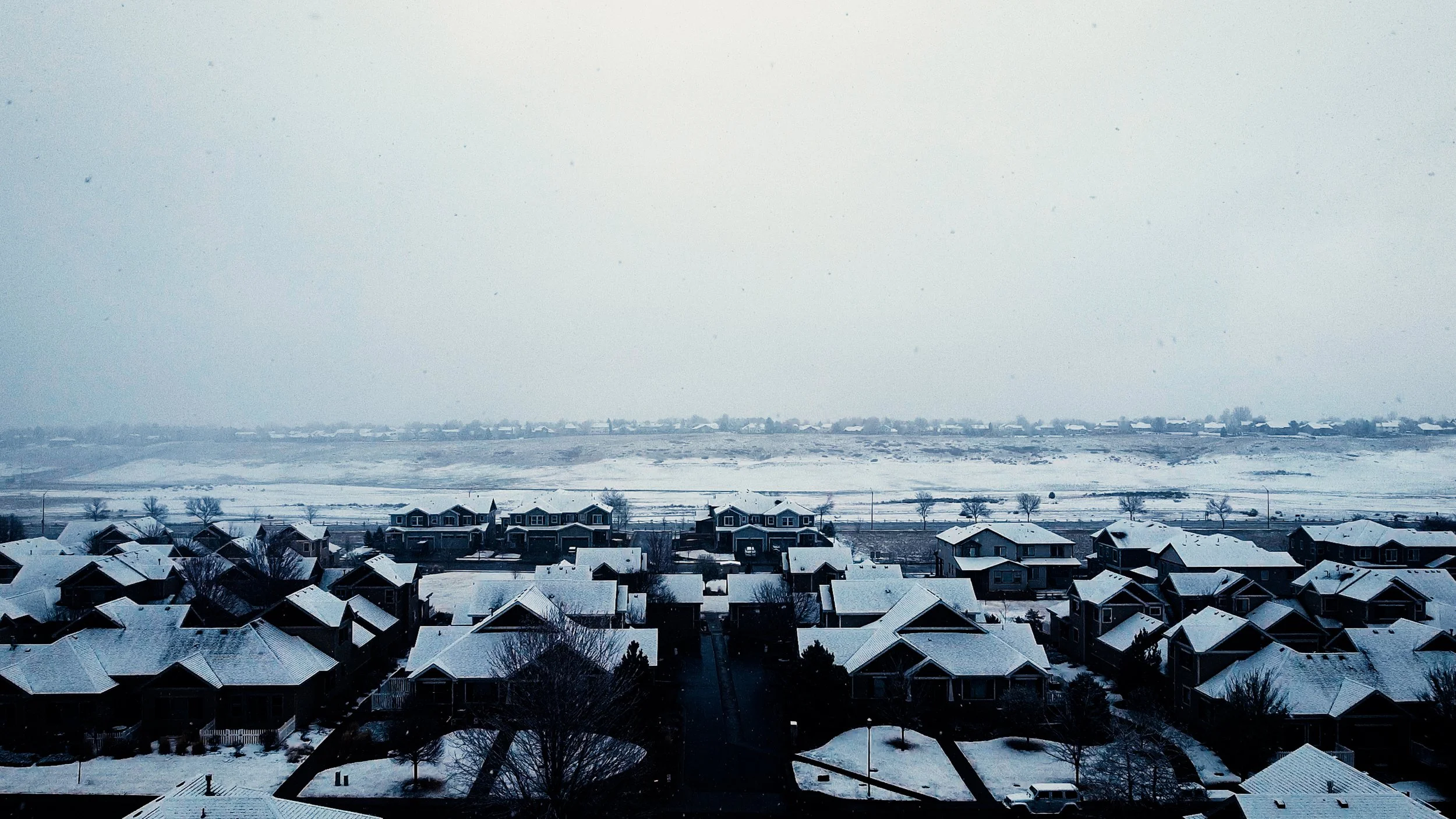 A snowy residential neighborhood with rooftops covered in snow, a few leafless trees, and a distant view of a snow-covered landscape under a cloudy sky.