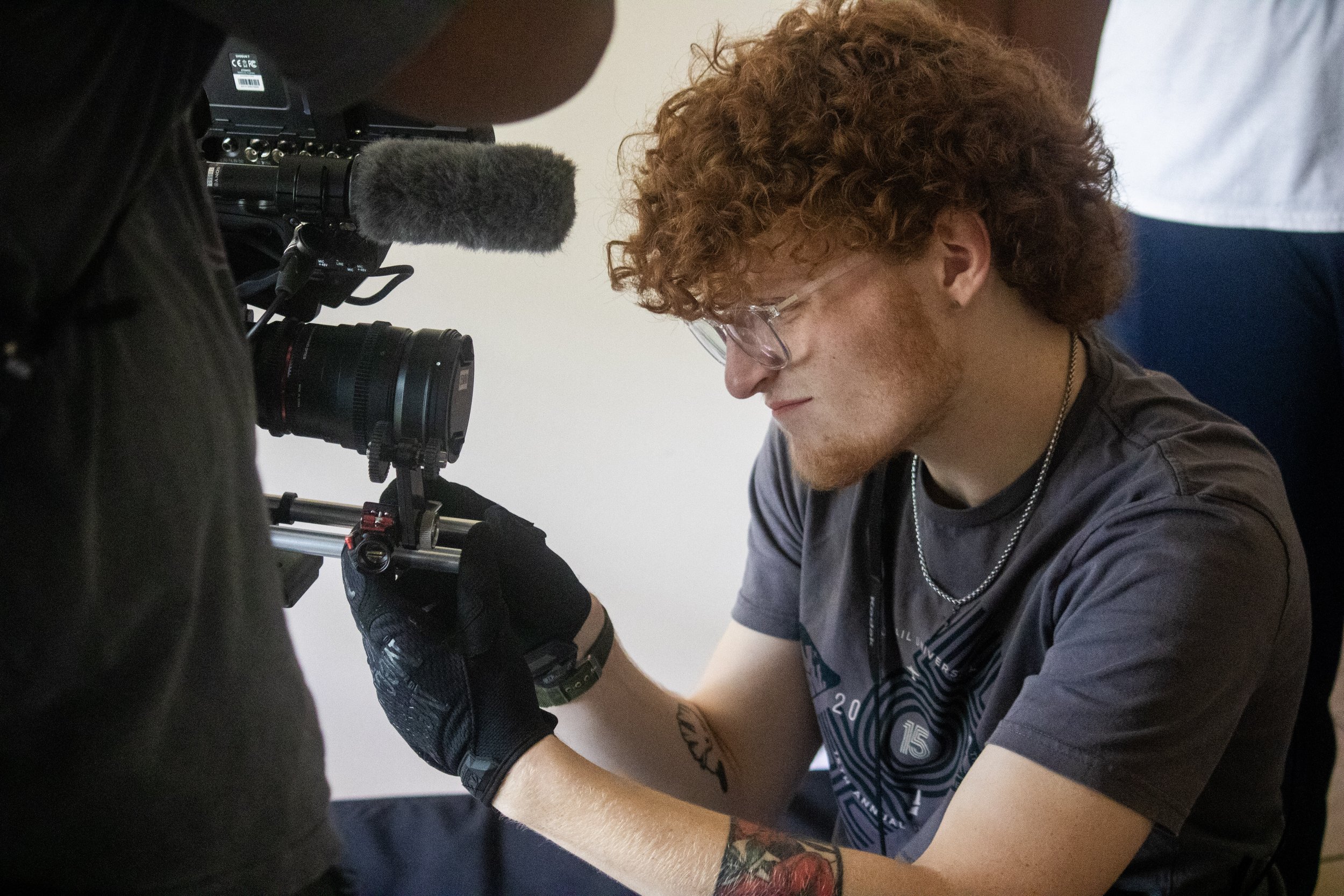 A person with curly red hair and glasses is getting a tattoo on their arm by a tattoo artist. The tattoo artist is using a tattoo machine and wearing black gloves.