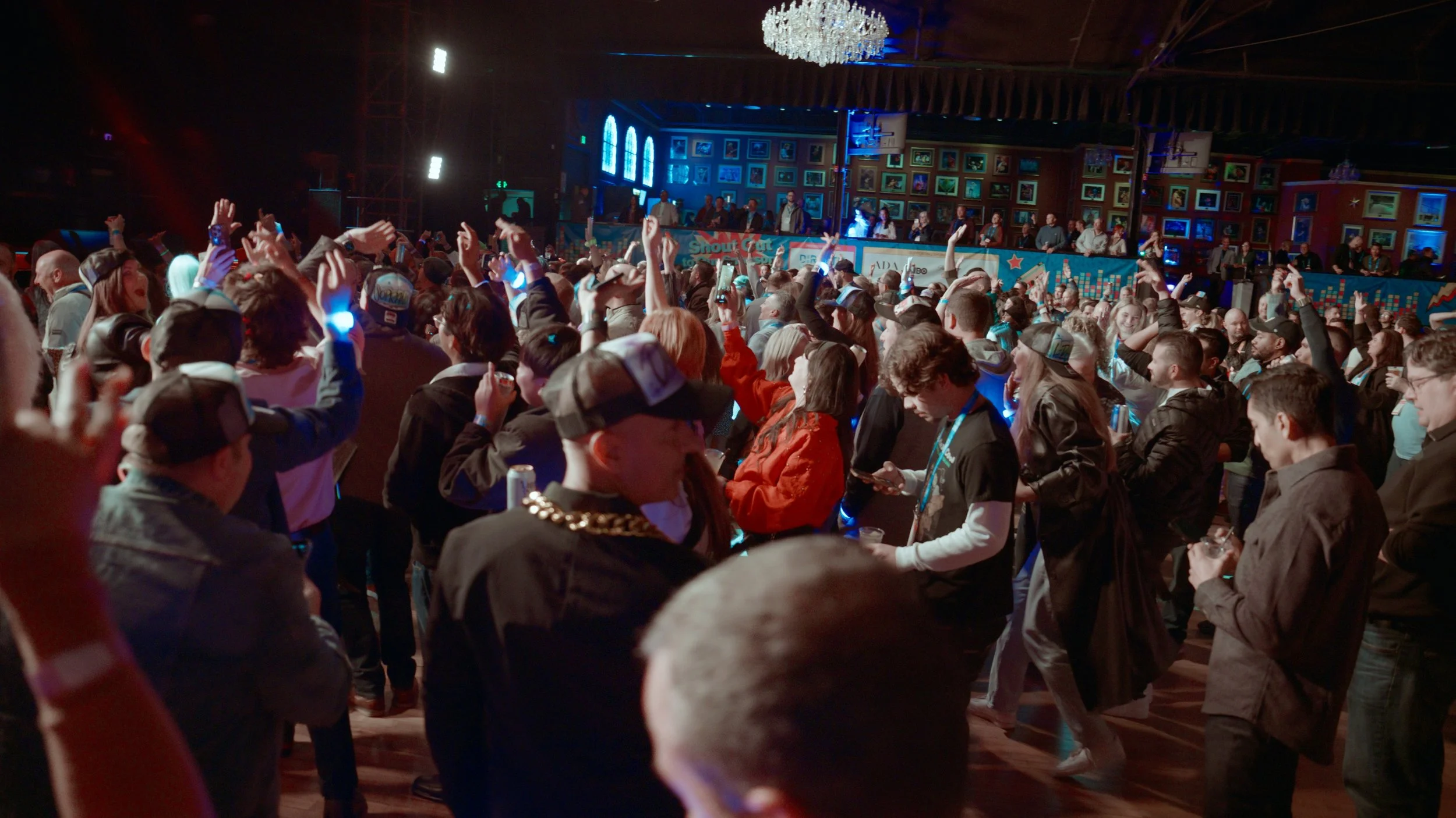 Crowd of people at a lively indoor event or concert, with some raising their hands and others using their phones, in a decorated venue with framed pictures on the wall and a chandelier overhead.