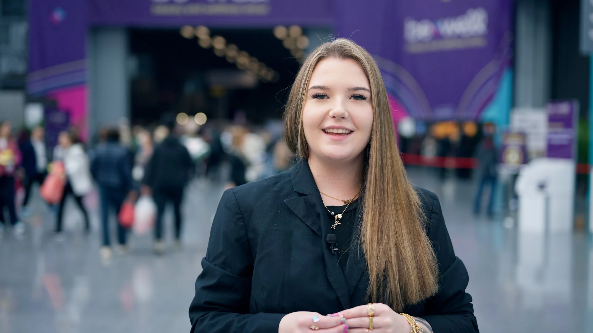 A young woman with long, straight hair smiling at the camera in an indoor public space with blurred people and purple signage in the background.