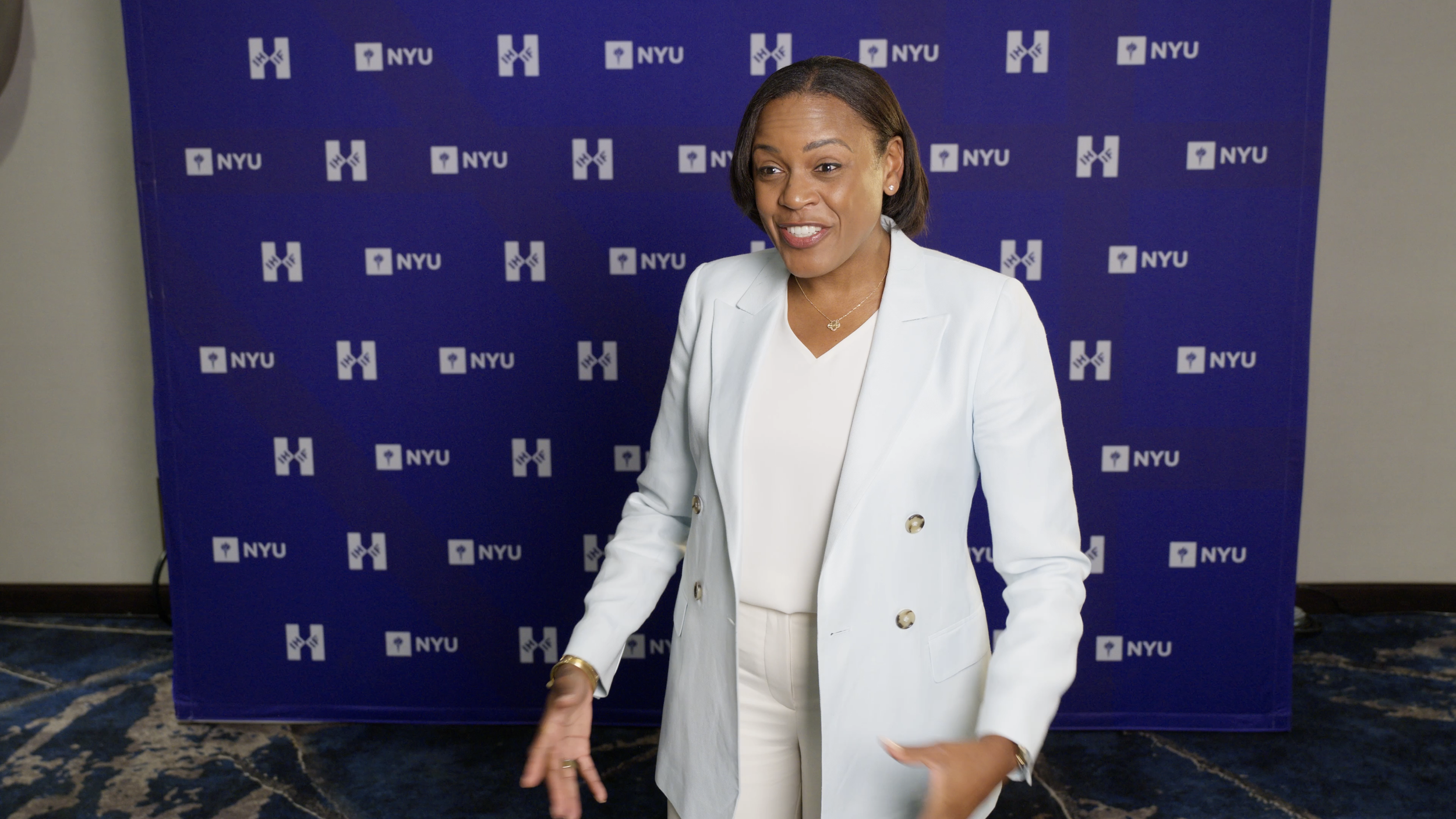 A woman in a white blazer standing in front of a blue NYU backdrop.