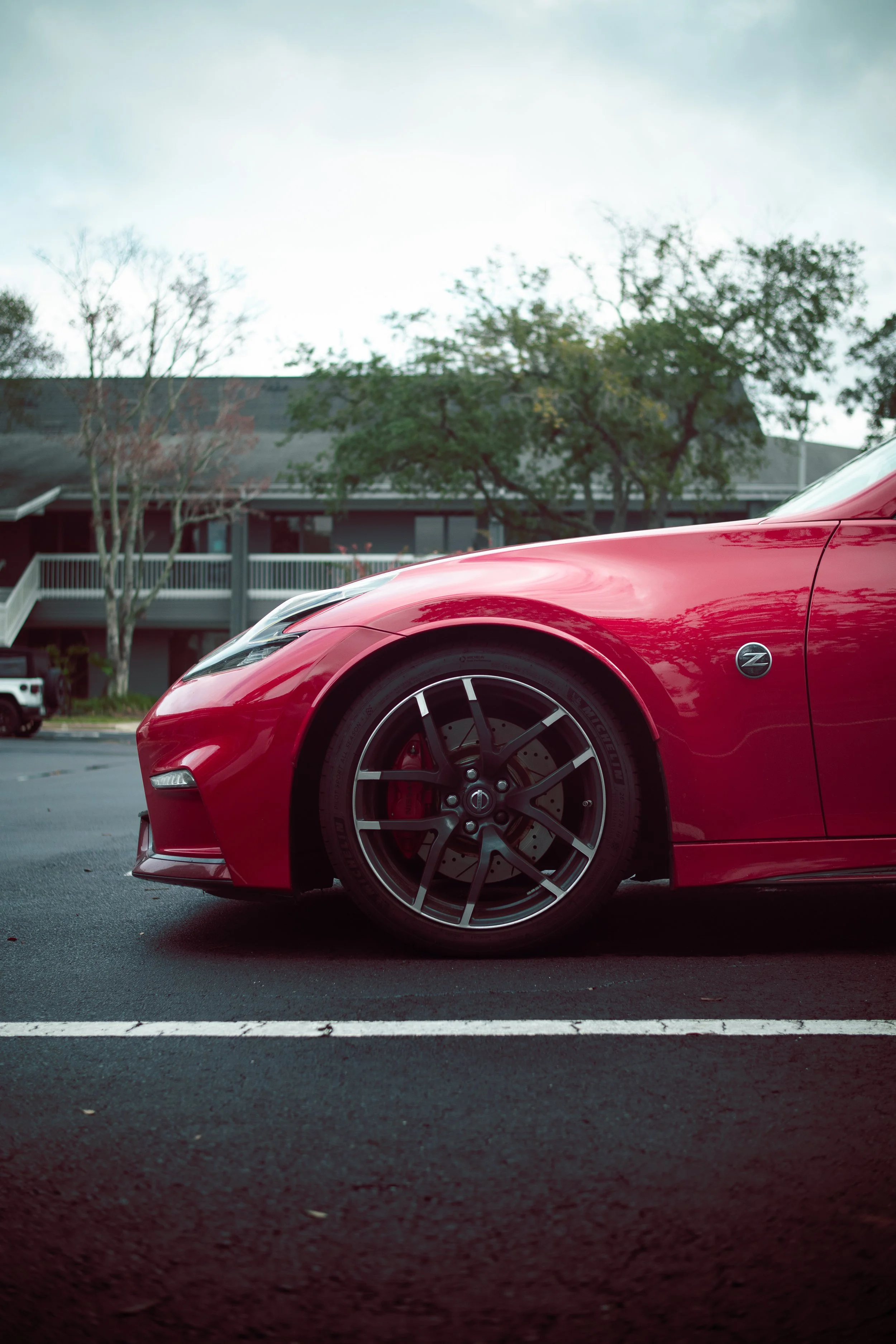 Close-up of a red sports car parked on a paved lot, with trees and a building in the background under an overcast sky.
