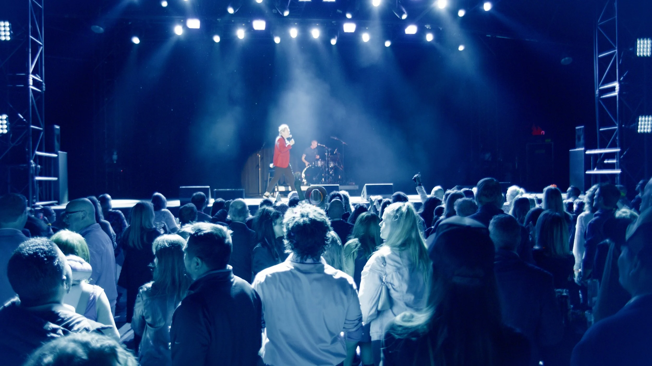 A singer wearing a red jacket performing on a stage with a drummer in the background at a concert, with a large audience watching.