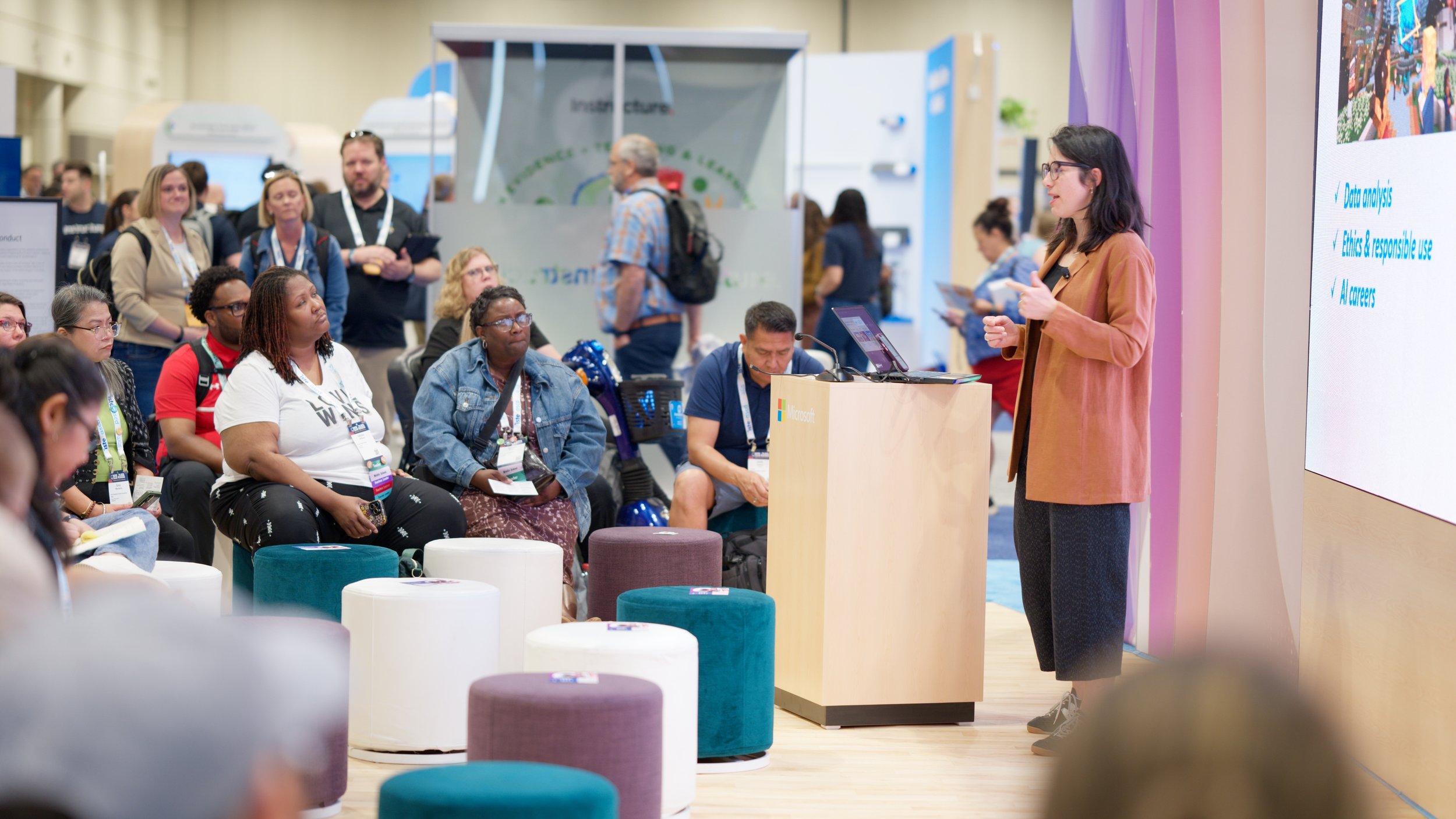 A woman wearing glasses and a brown blazer is giving a presentation to a seated audience at a conference or expo. The audience includes diverse individuals taking notes or listening attentively. A large screen with a presentation slide is visible bes