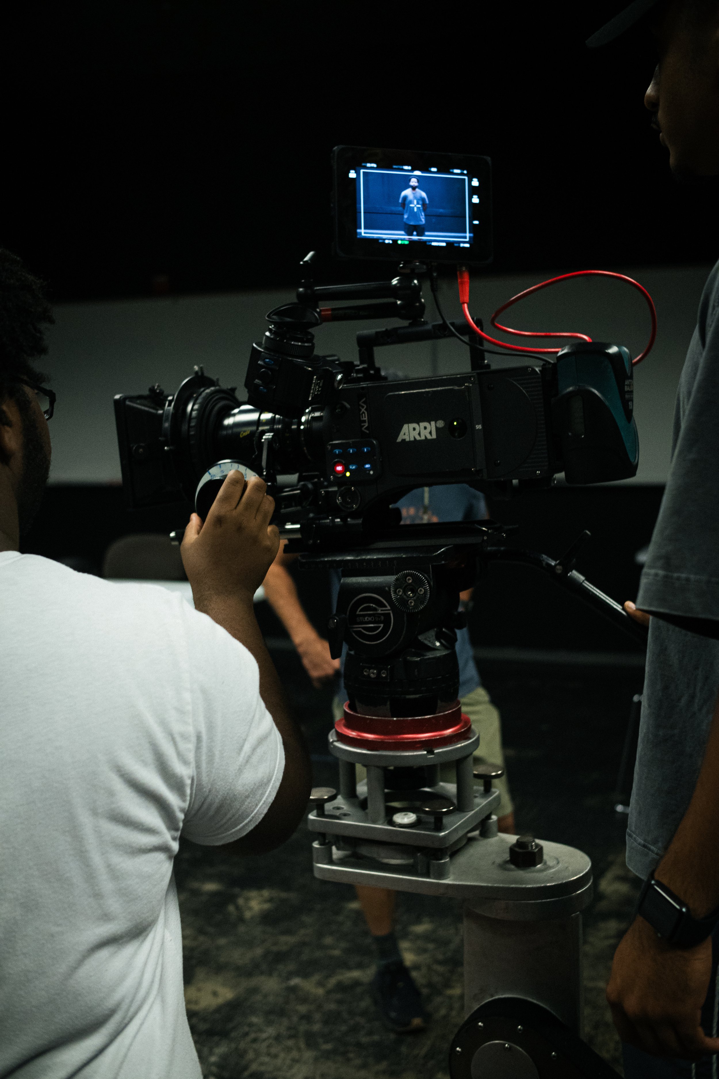 Professional camera setup filming a person standing against a dark background, with a monitor displaying the person being filmed.