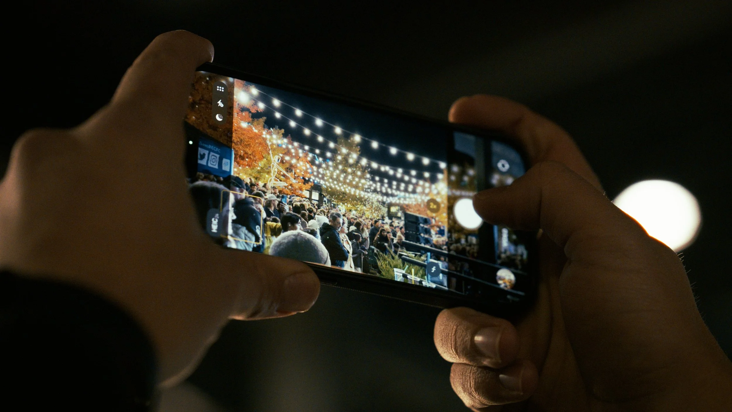 A person holding a smartphone captures a photo of an outdoor festival or event decorated with string lights and lit-up trees at night, with a crowd of people in the background.